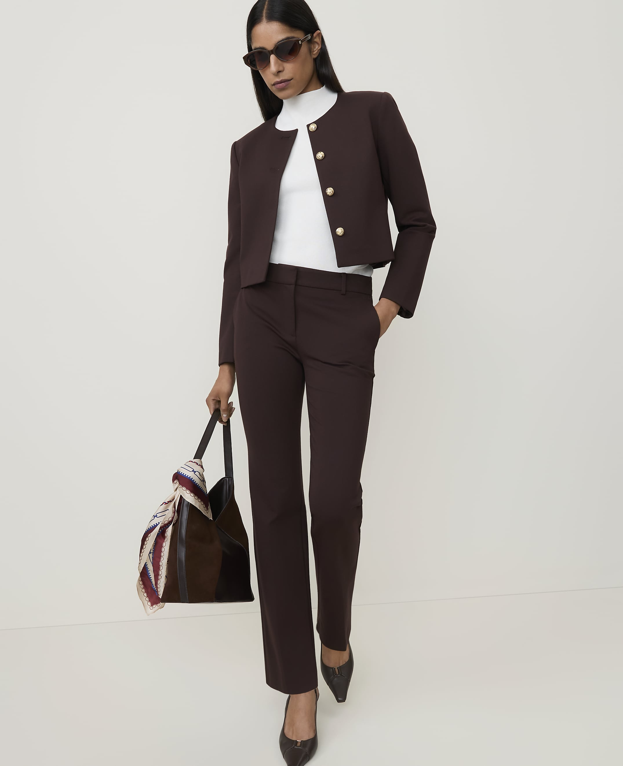 A woman in a brown suit and white turtleneck stands with a patterned scarf draped over her arm against a plain white background.