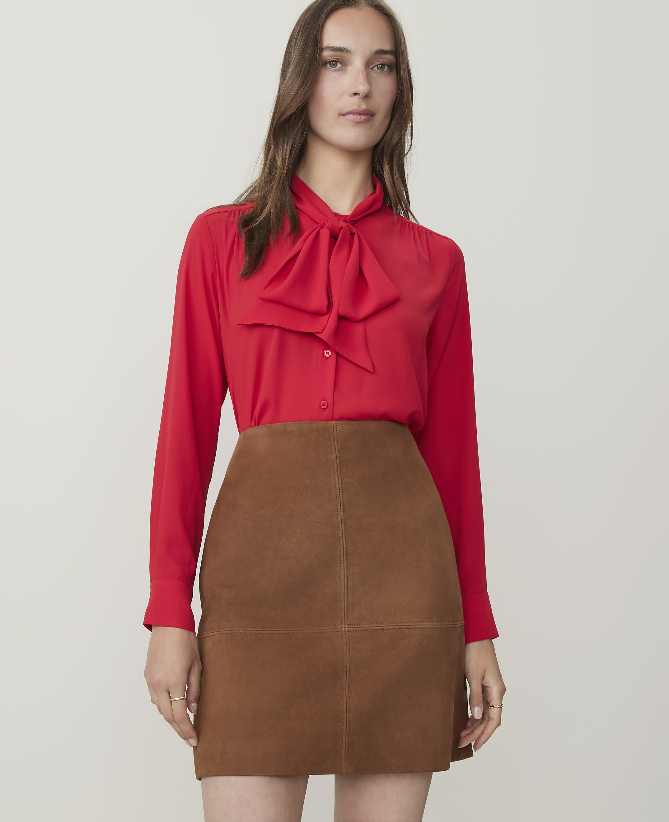 A woman in a red bow-neck blouse and brown suede skirt stands against a plain white background.