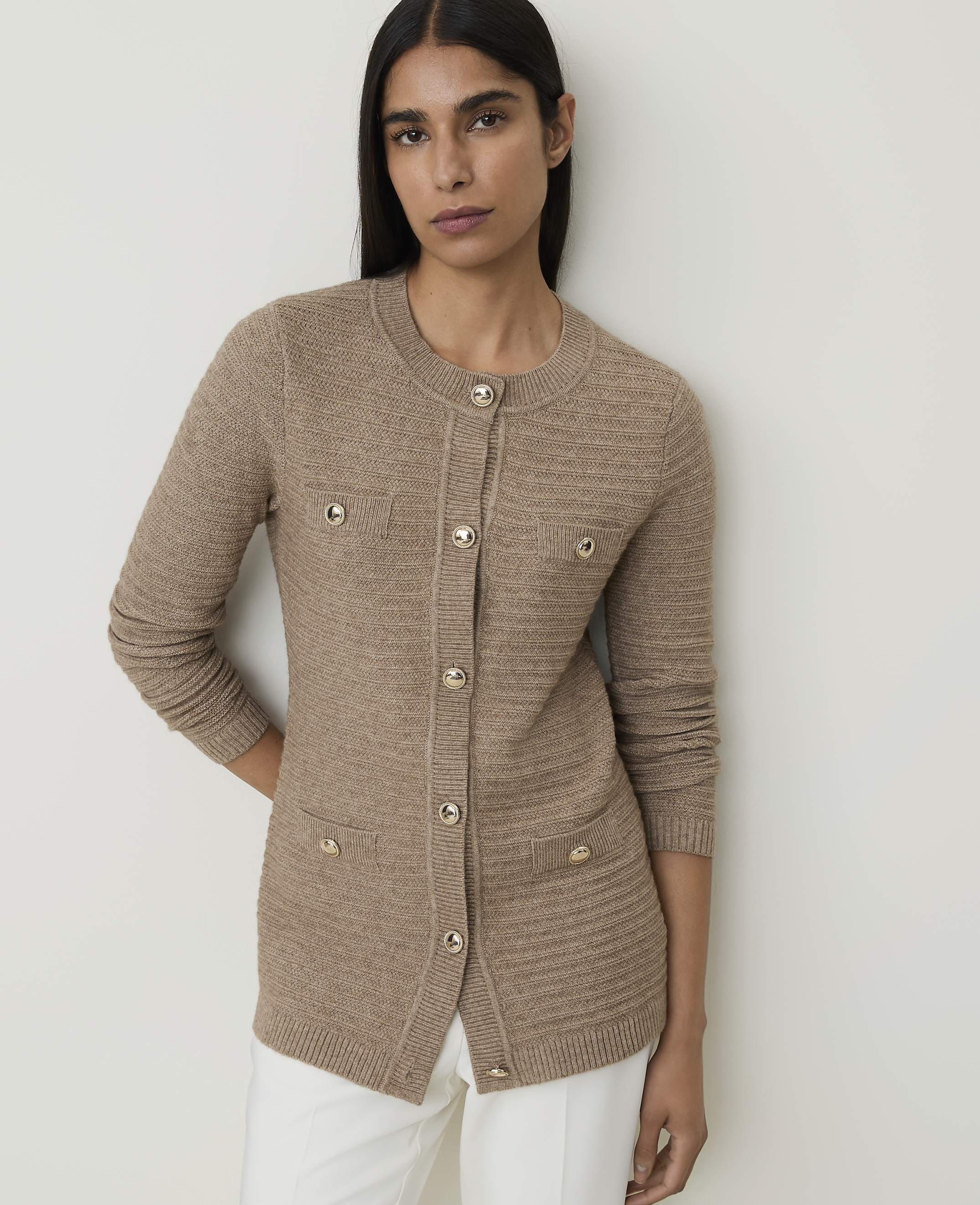 A woman in a beige cardigan and white pants stands against a plain, light gray background.