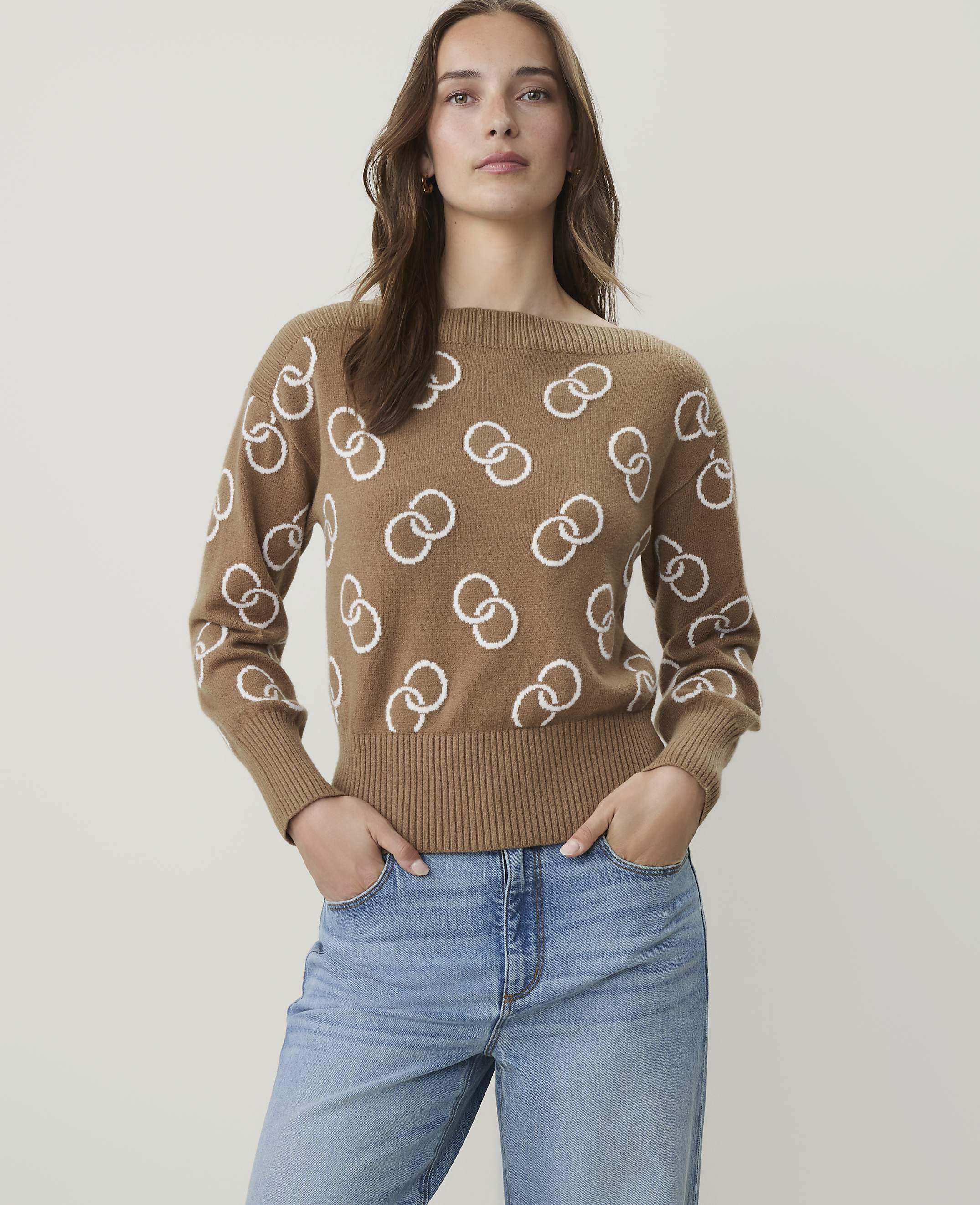 A woman in a tan sweater with a white interlocking ring pattern stands against a plain white background.