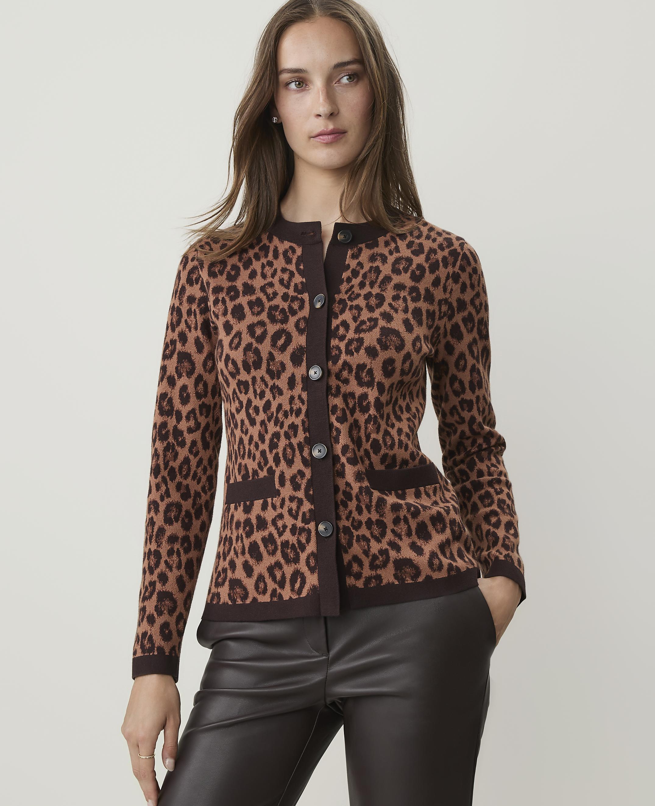 A woman in a leopard print cardigan and dark pants stands against a plain, light brown background.