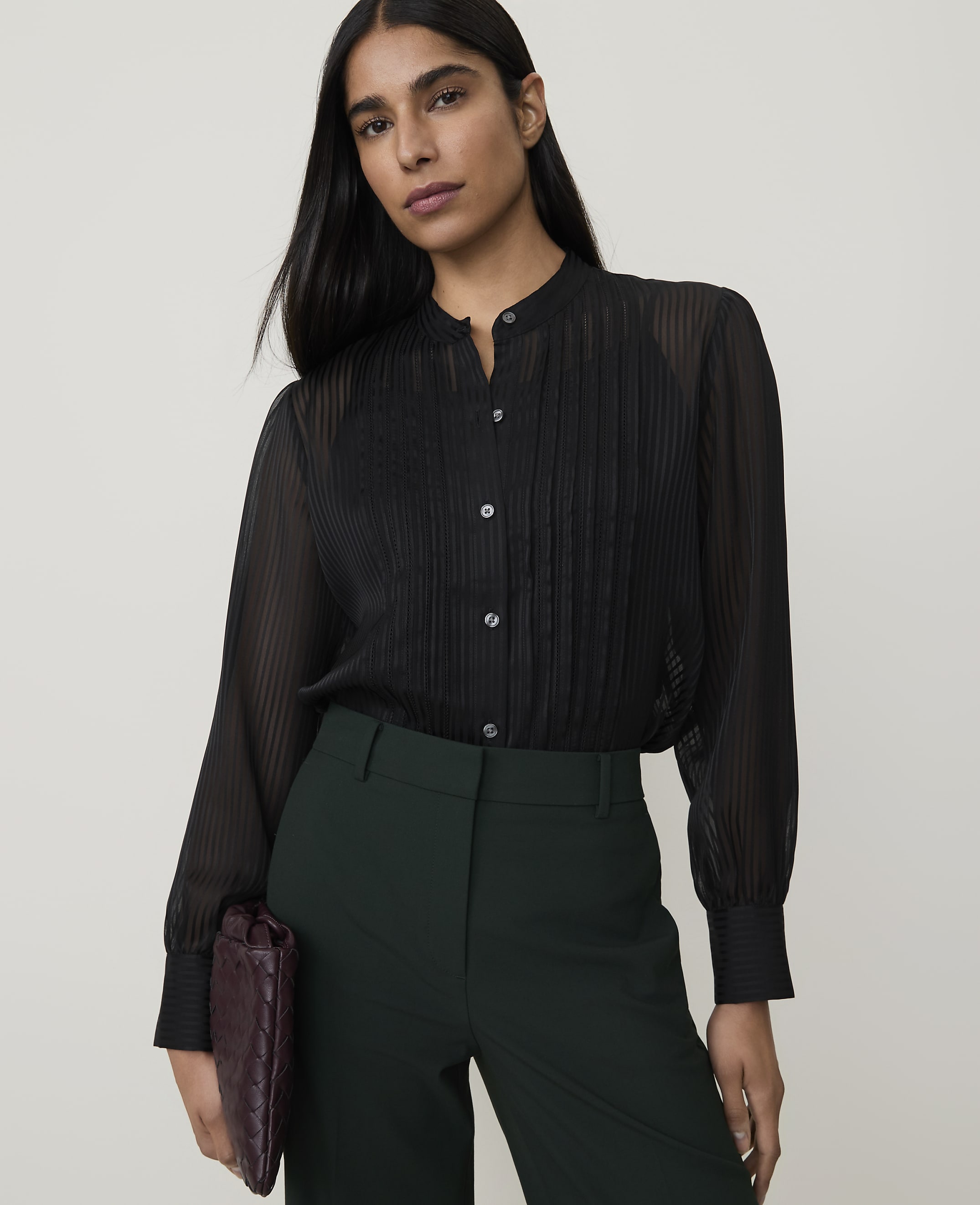 A woman in a sheer black blouse and dark green pants holds a burgundy clutch against a plain white background.