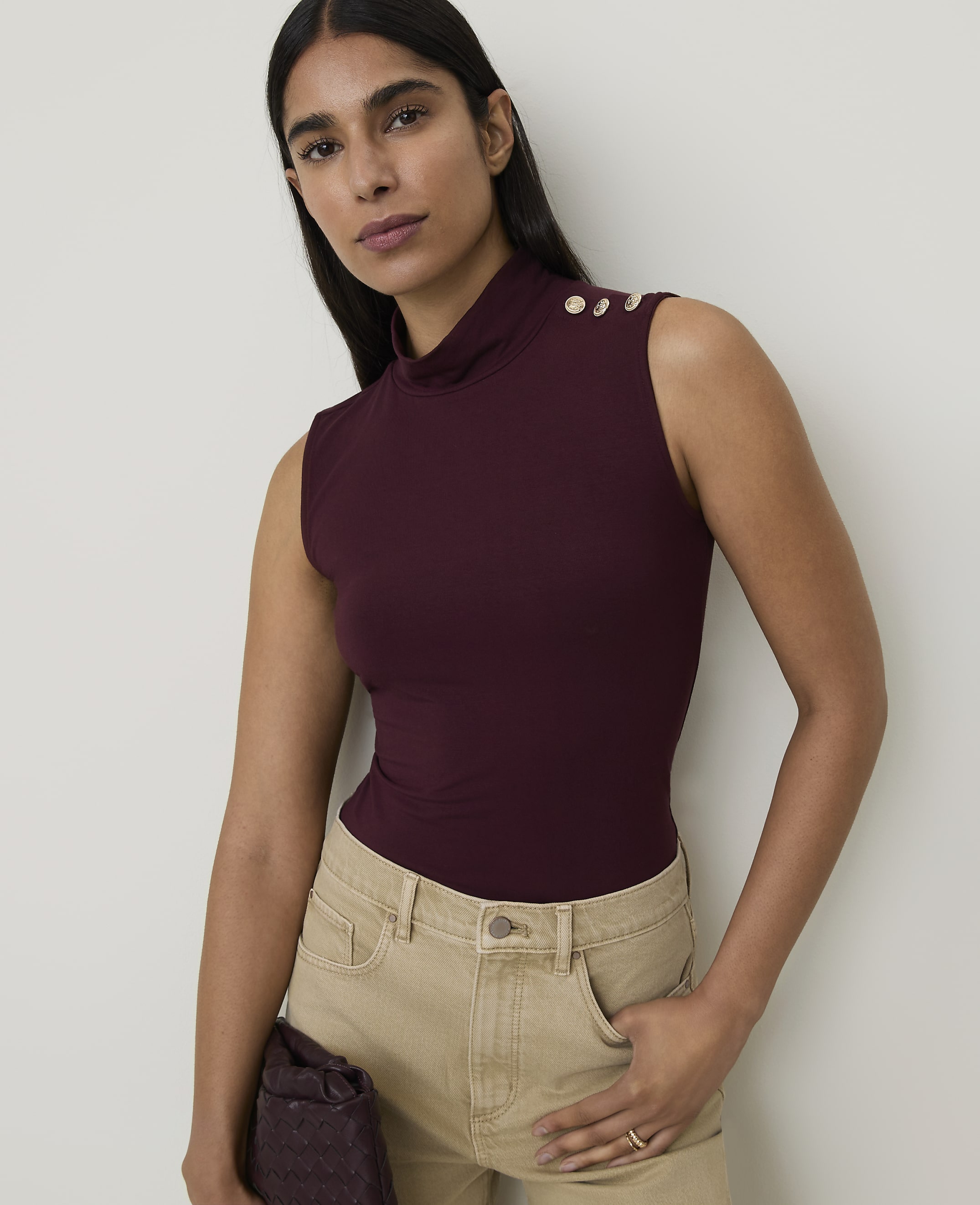 A woman in a burgundy turtleneck top and tan pants stands against a plain white background.