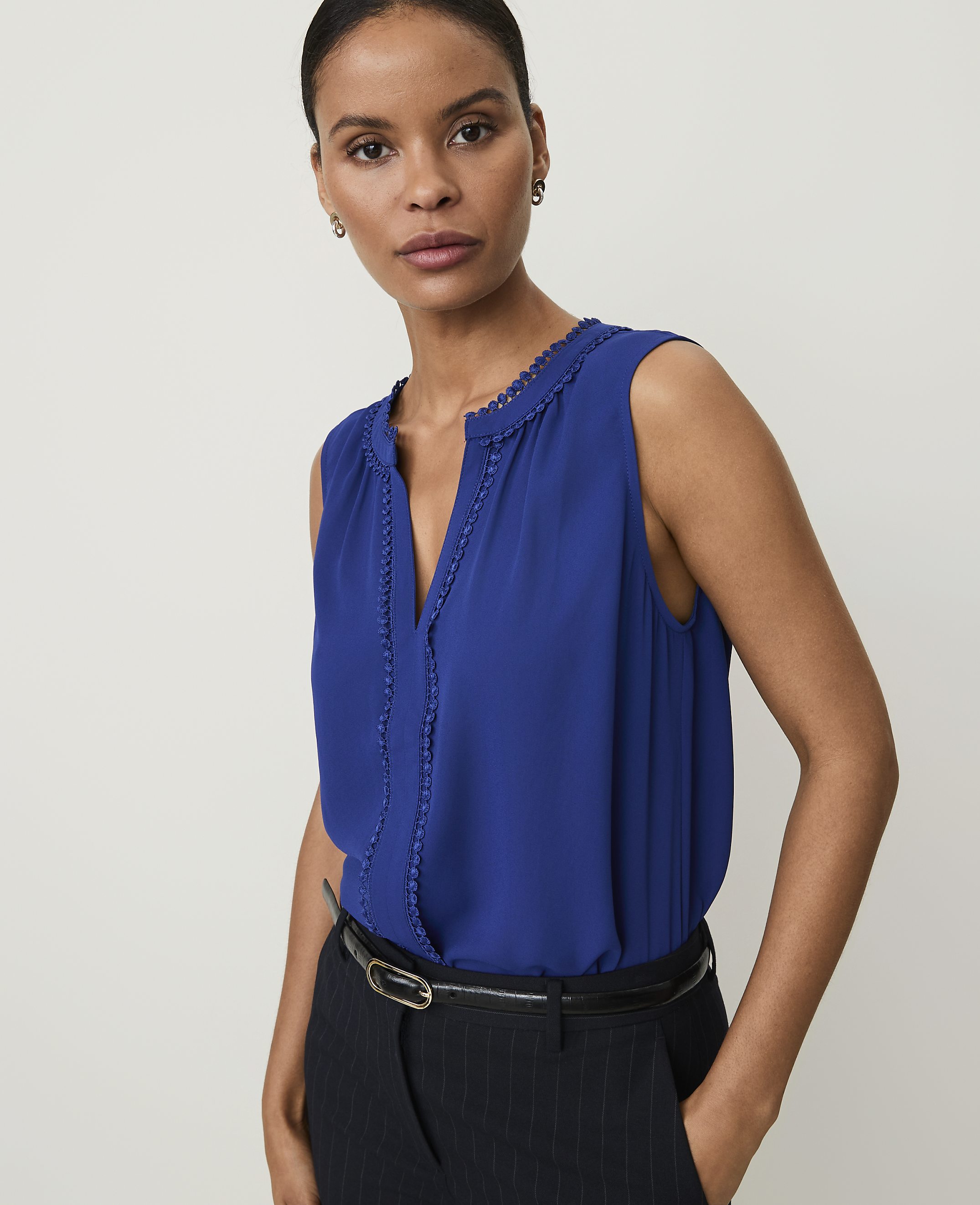 A woman in a blue sleeveless top and pinstripe pants stands against a plain white background.