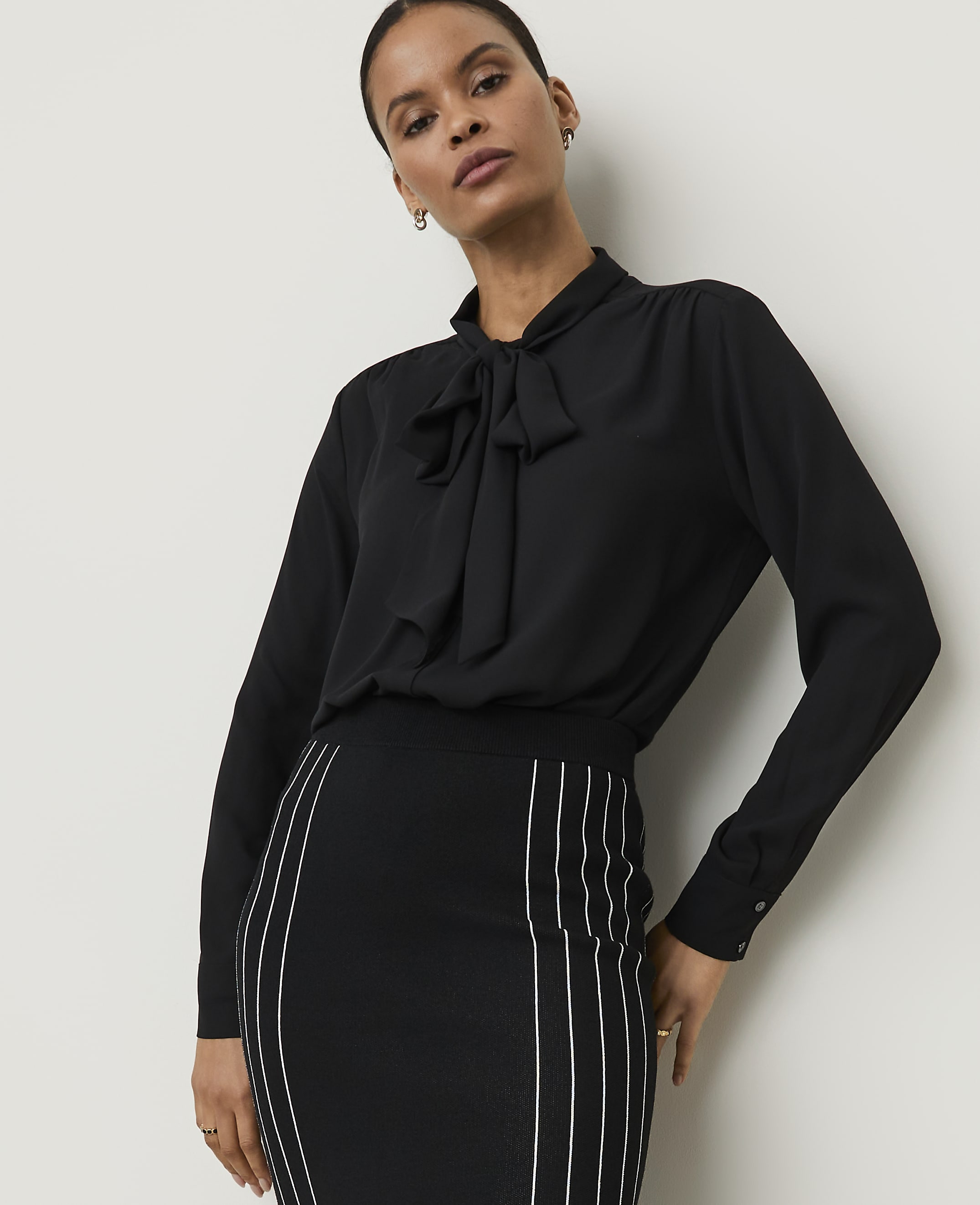 A woman in a black pussy-bow blouse and a striped pencil skirt stands against a plain white background.