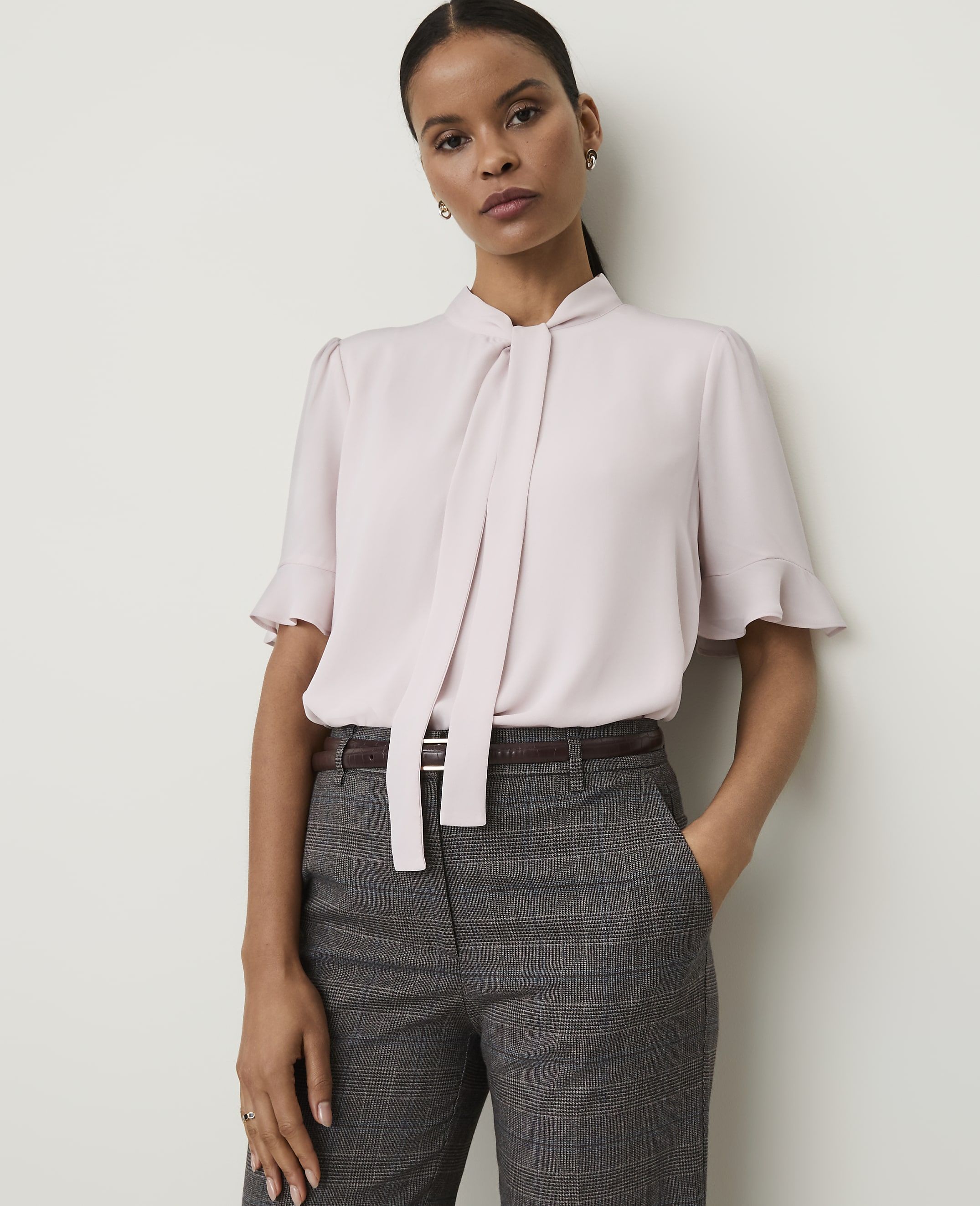 A woman in a light pink bow-neck blouse and plaid trousers stands against a plain white background.