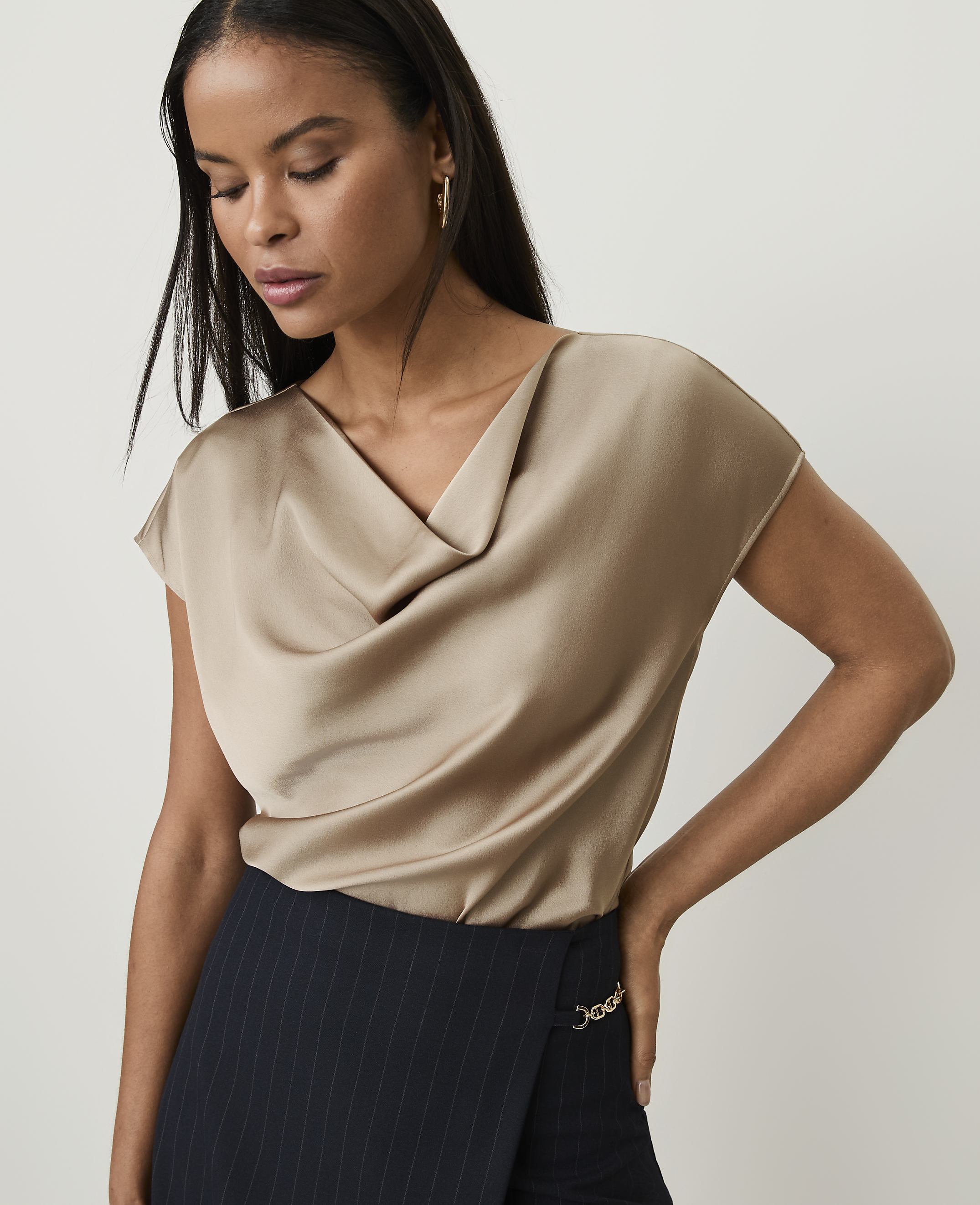 A woman in a champagne-colored cowl-neck top and a pinstripe skirt stands against a plain, light background.