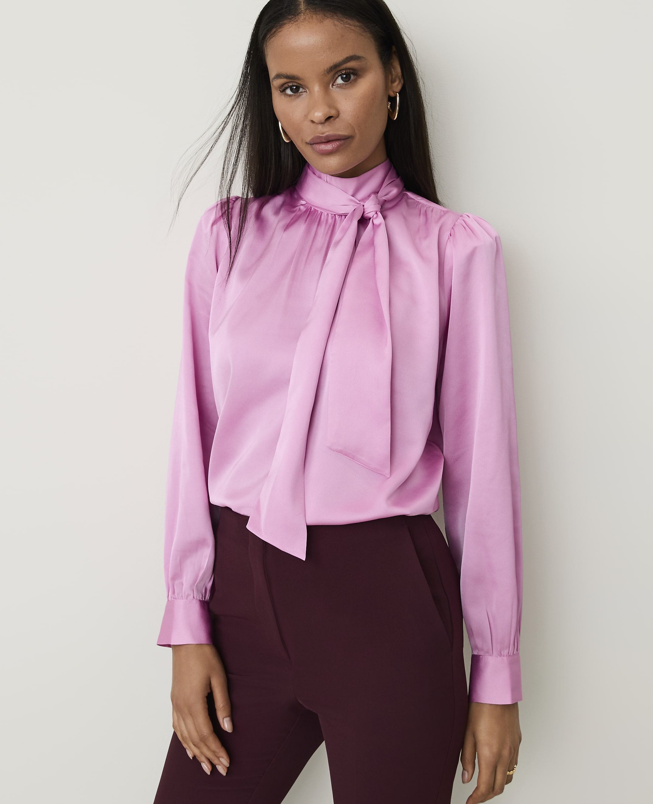 A woman in a pink satin blouse and burgundy pants stands against a plain white background.
