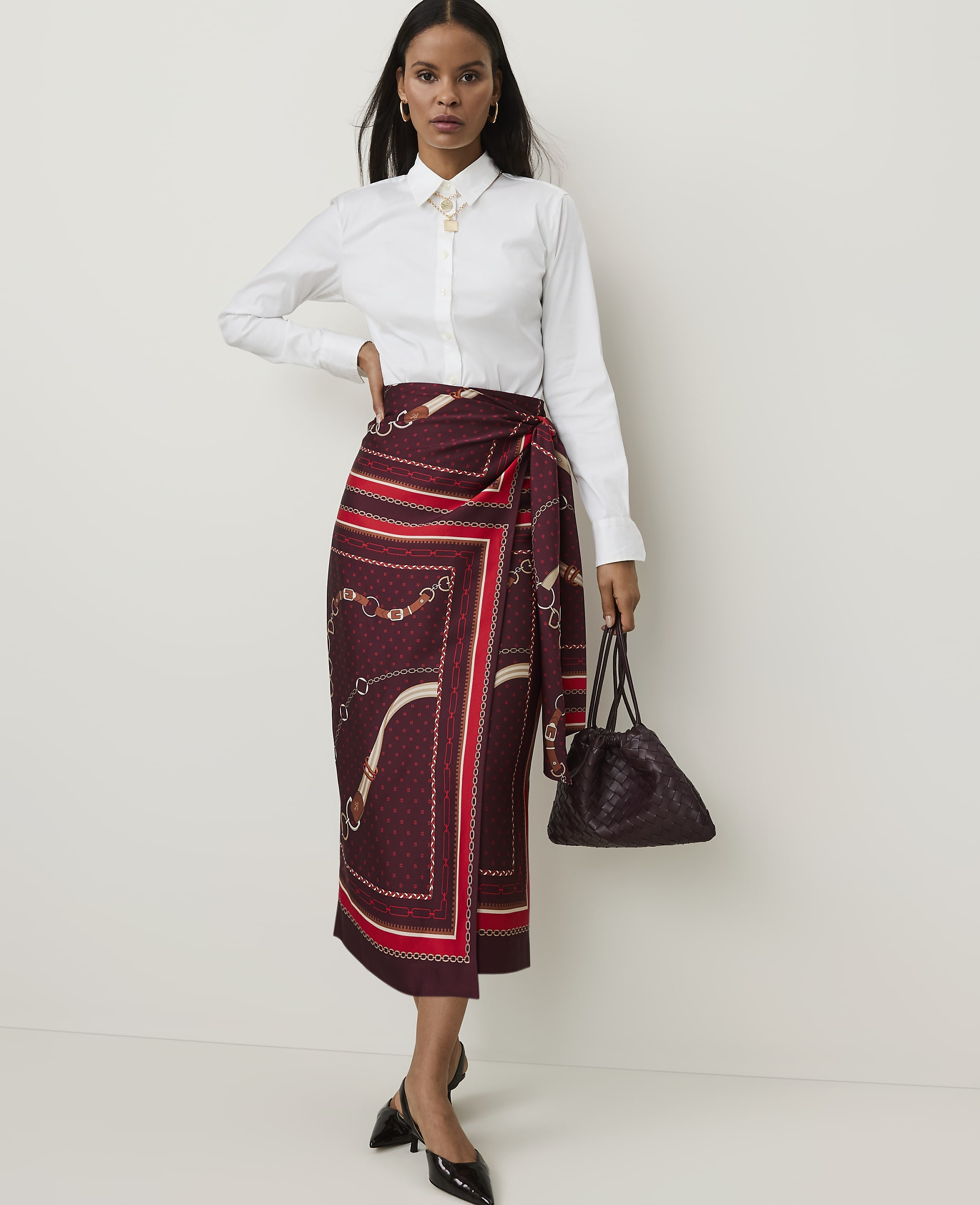 A woman in a white collared shirt and a patterned wrap skirt stands against a plain white background, holding a dark handbag.