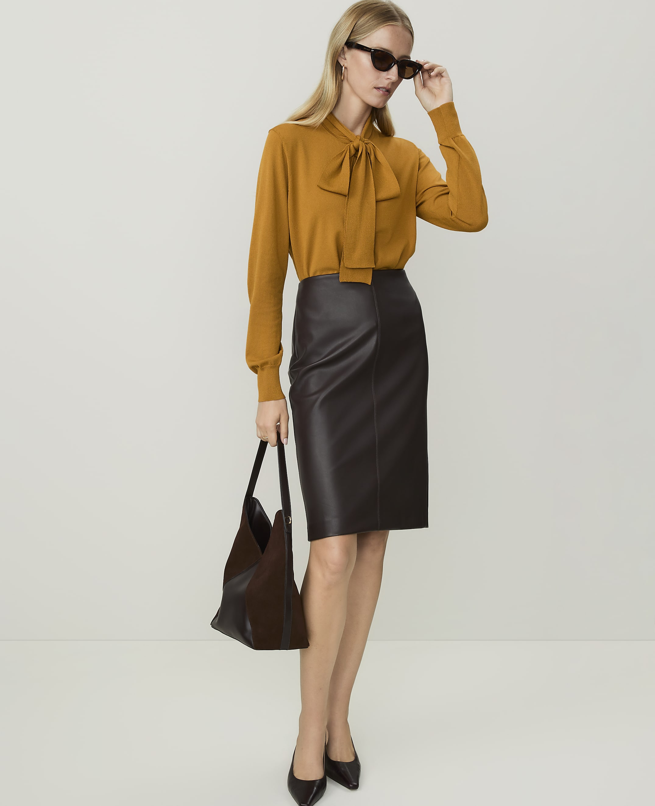 A woman in a mustard-colored bow-neck sweater and a dark brown leather pencil skirt stands with a dark handbag against a plain white background.