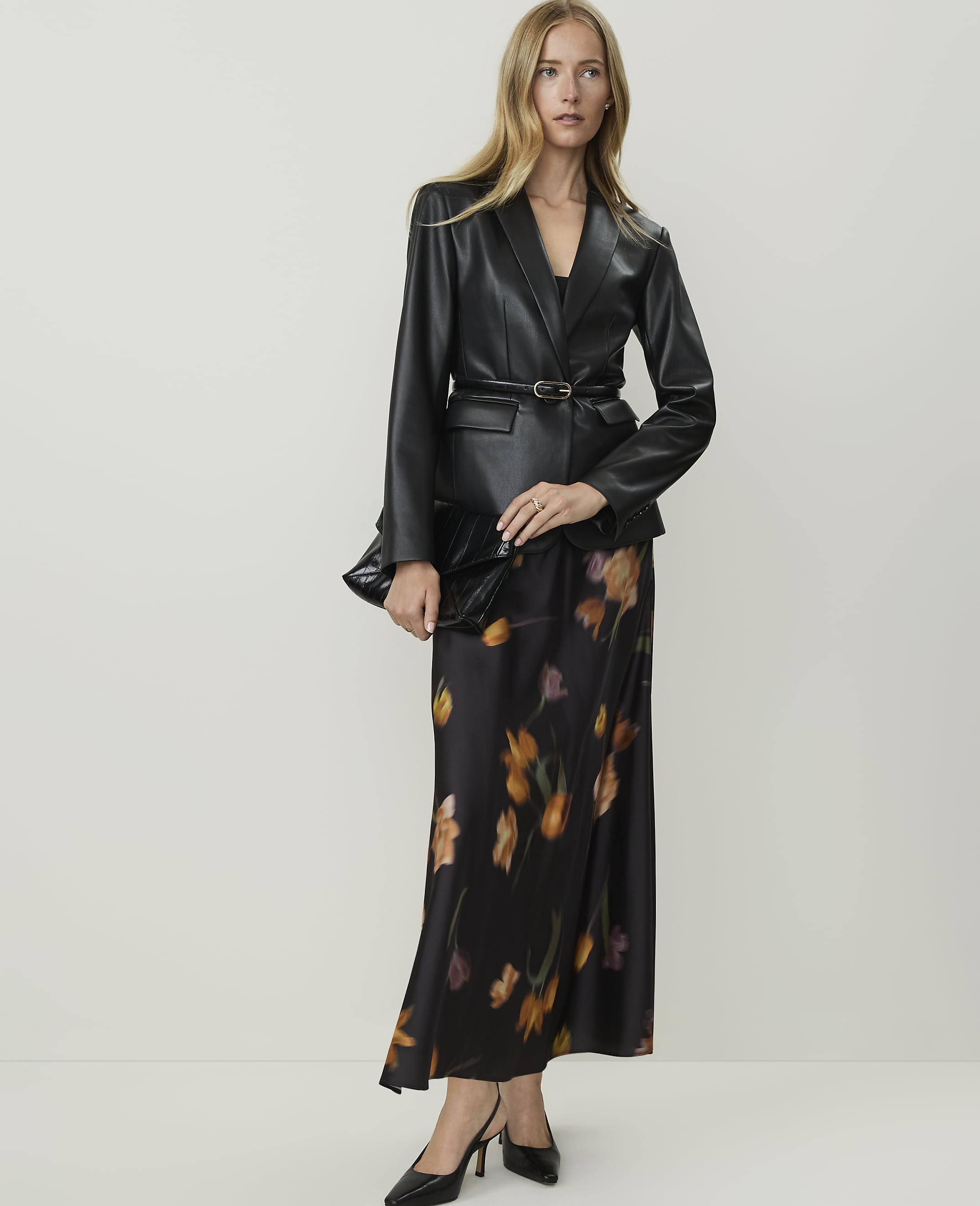 A woman in a black leather blazer and a floral print skirt stands against a plain white background.