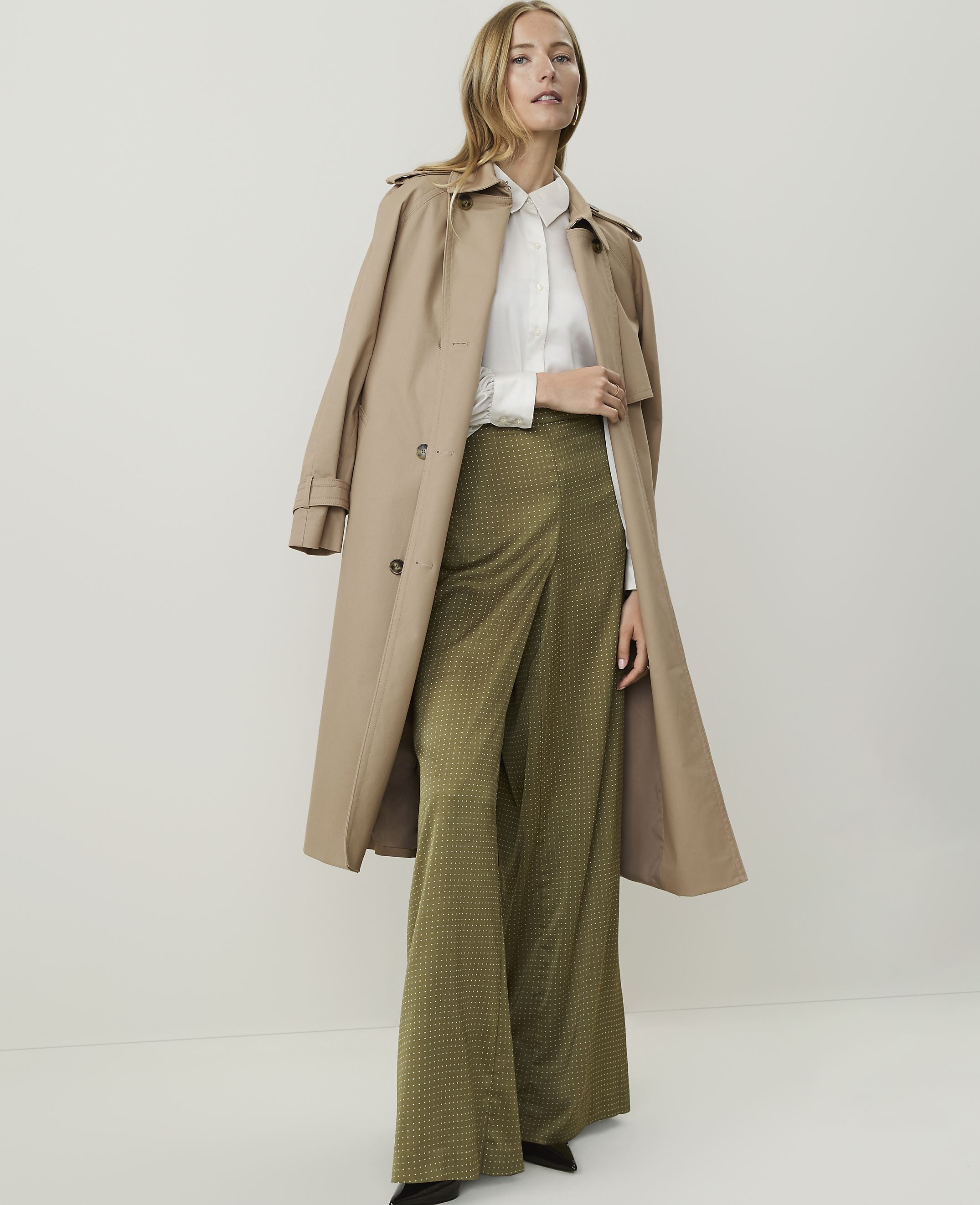 A woman in a trench coat and patterned pants stands against a plain, light background.