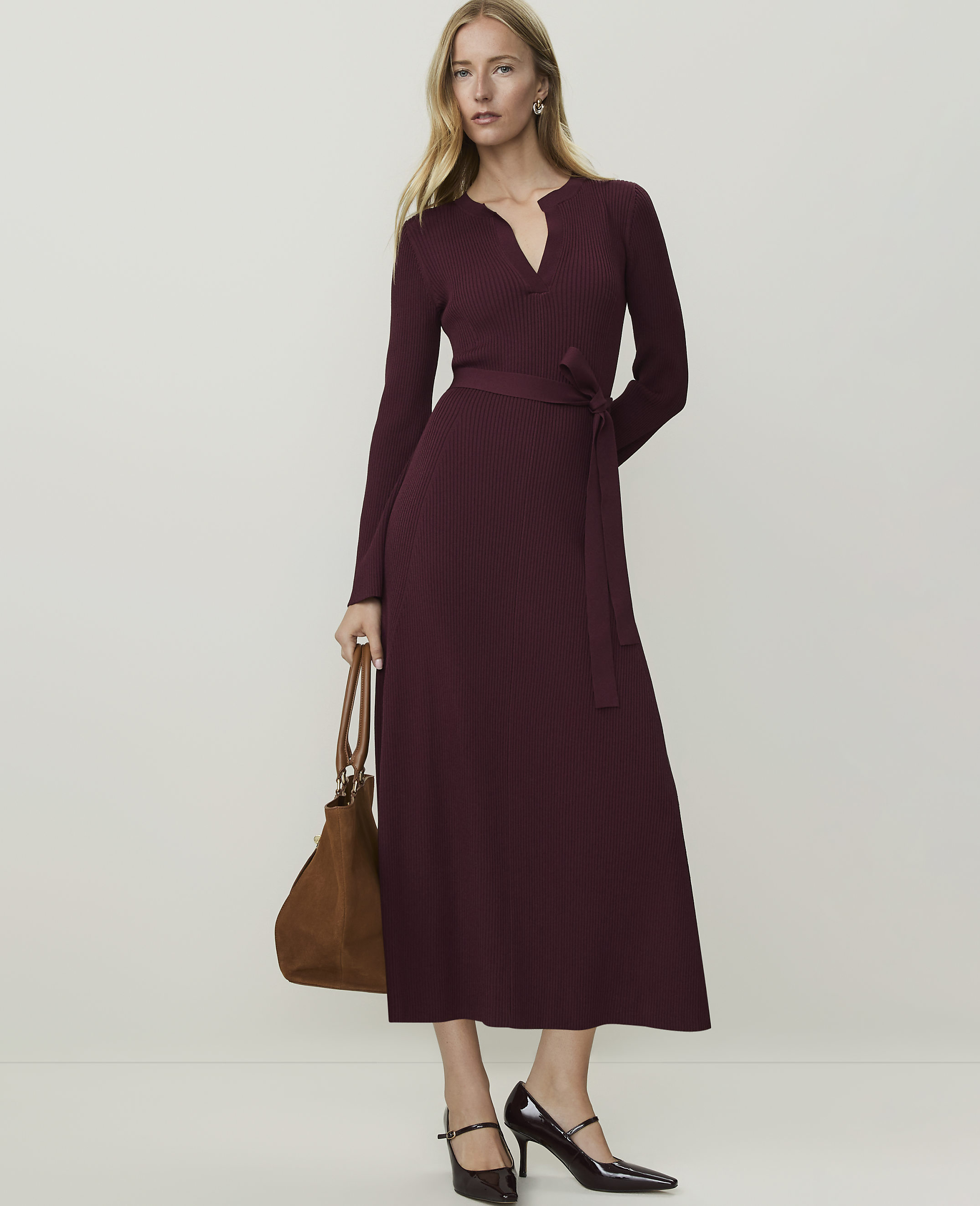 A woman in a burgundy ribbed dress and heels stands with a brown bag against a plain, light background.