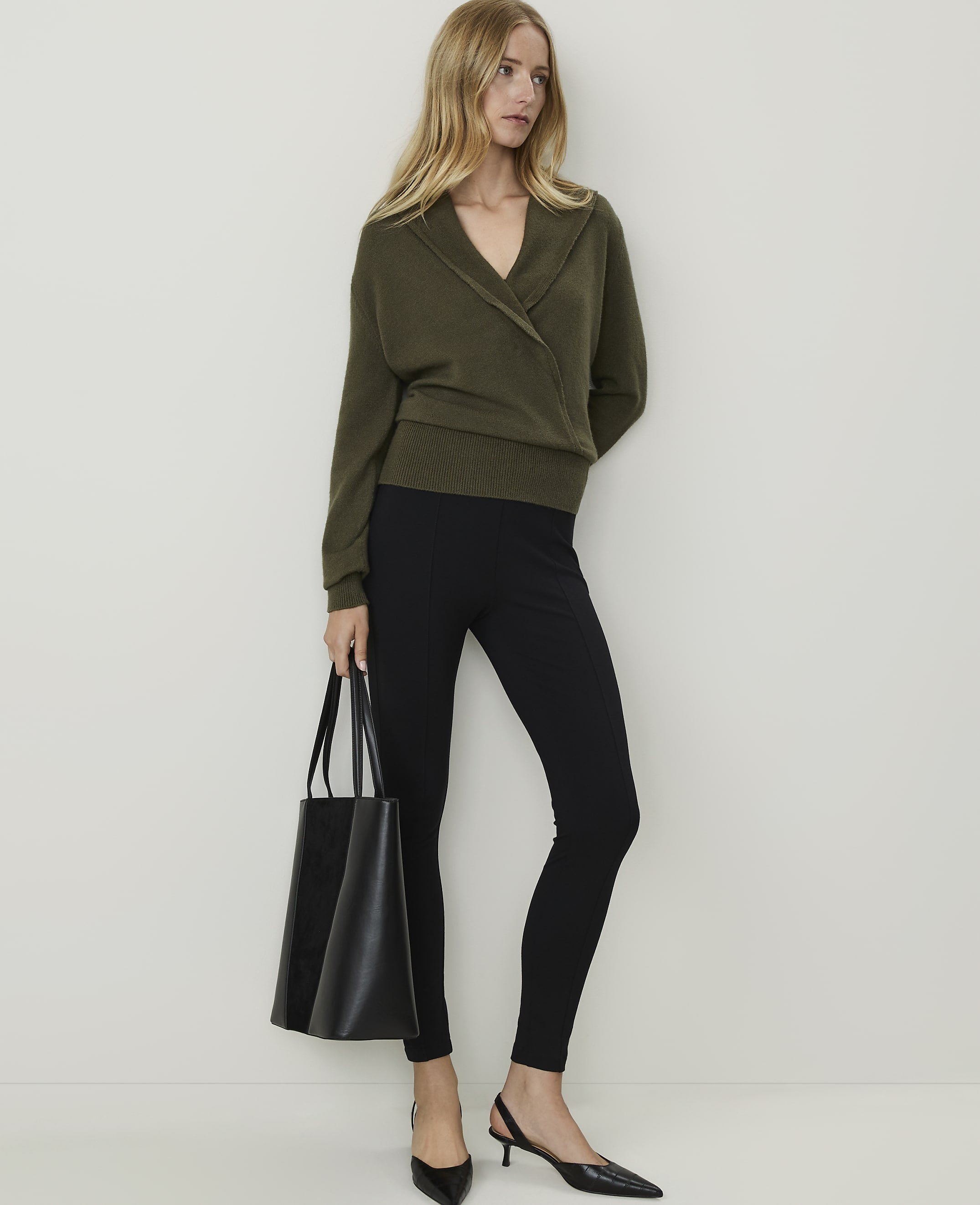 A woman in a green wrap sweater and black leggings stands with a black tote bag against a plain white background.
