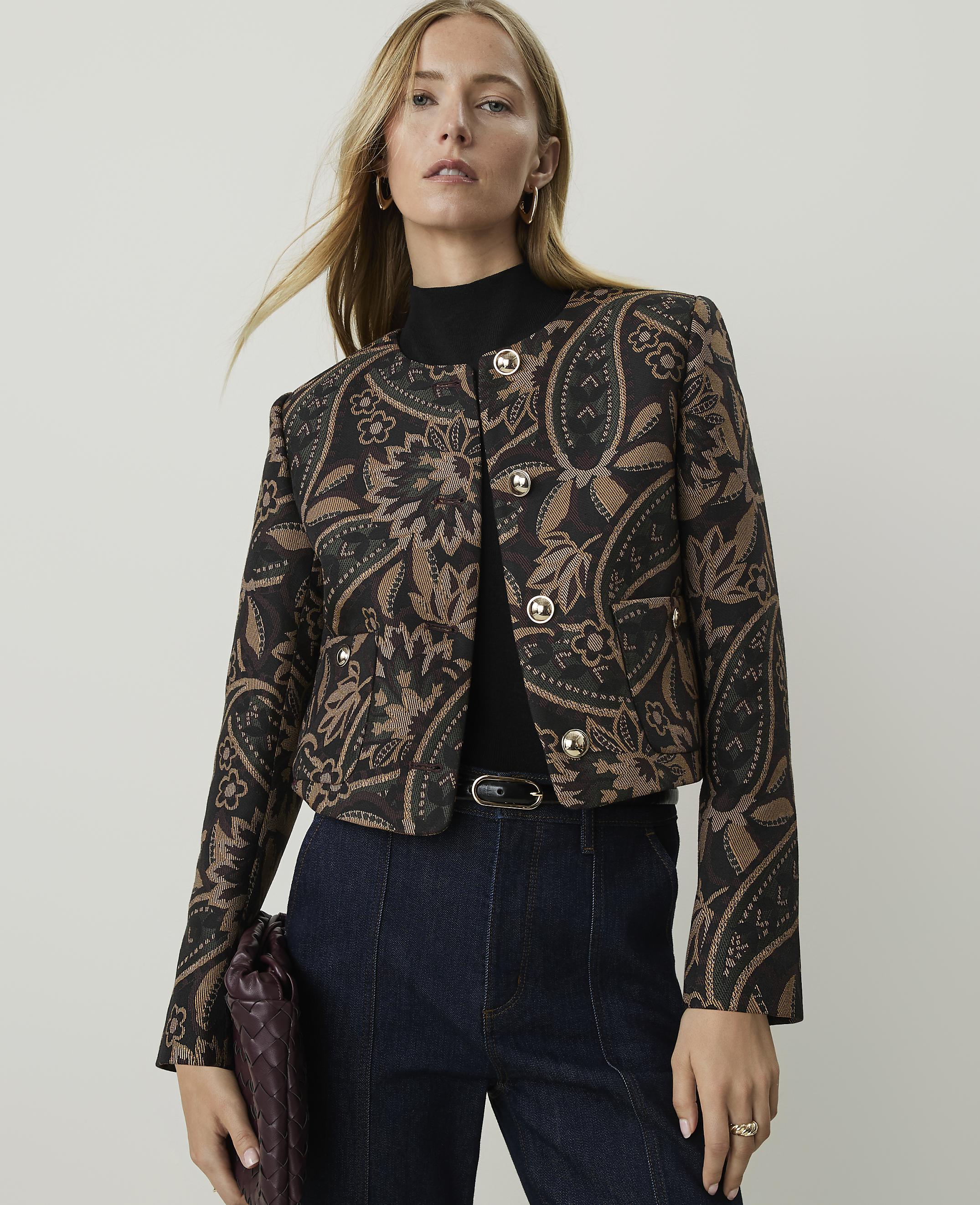 A woman in a patterned jacket and dark jeans stands against a plain, light background.