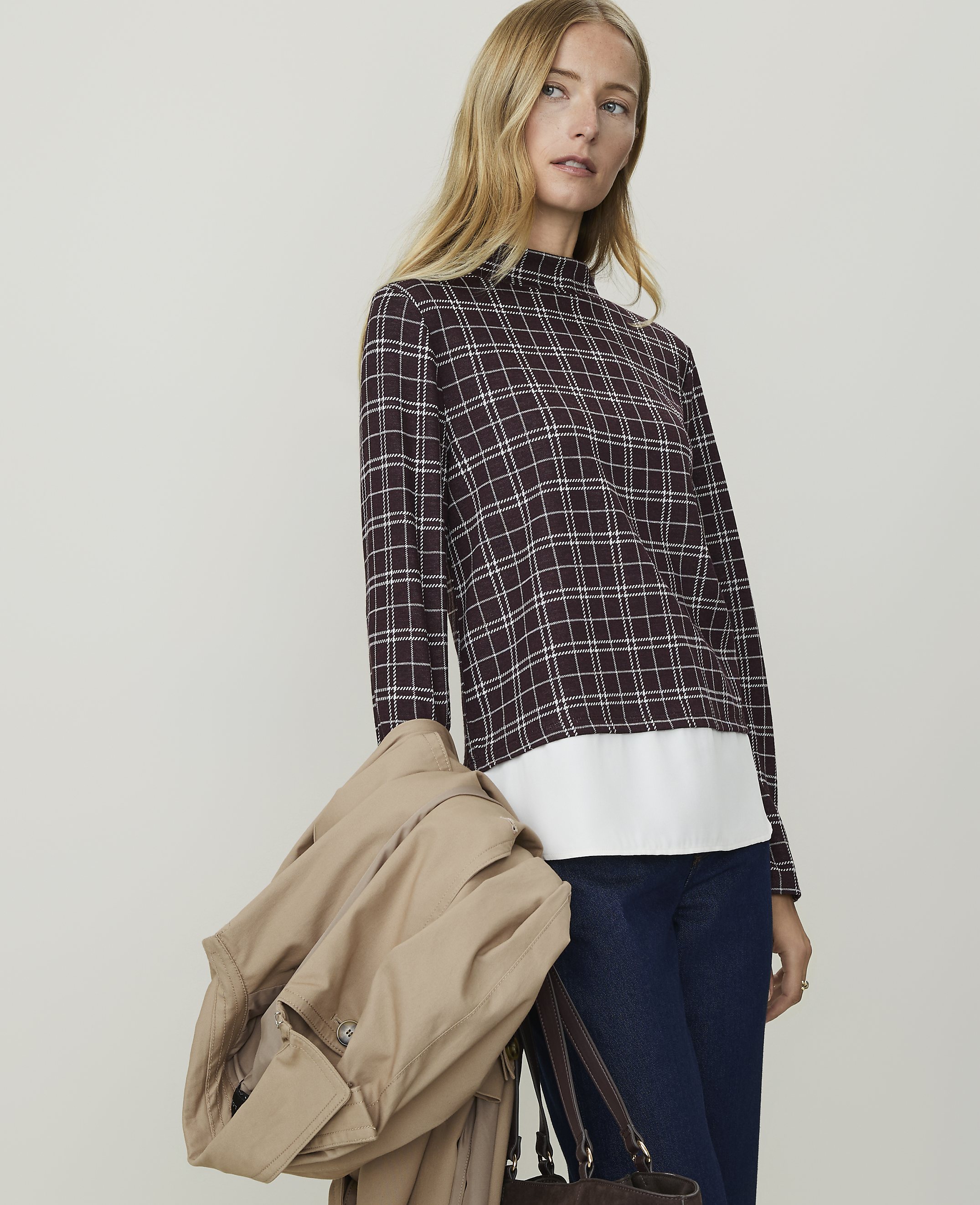 A woman in a plaid top and jeans carries a tan coat and a brown bag against a plain background.