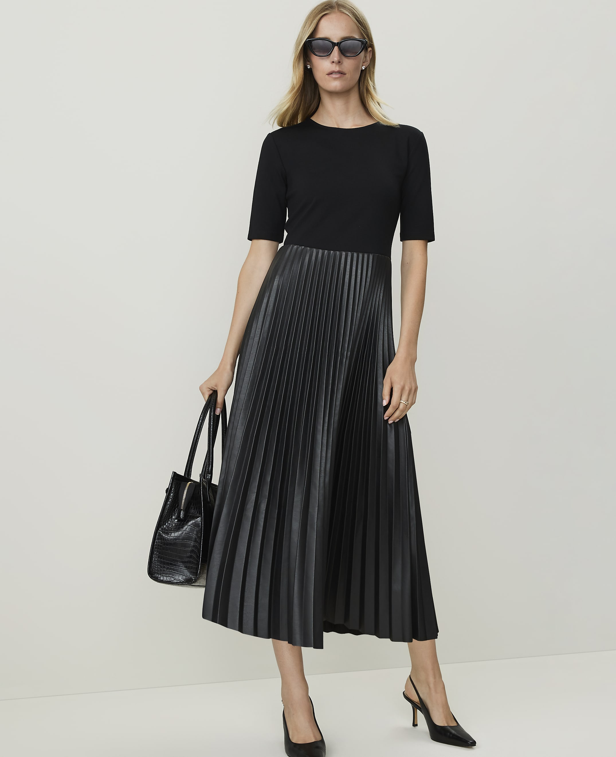 A woman in a black top and pleated faux leather skirt stands against a plain white background, holding a black handbag.