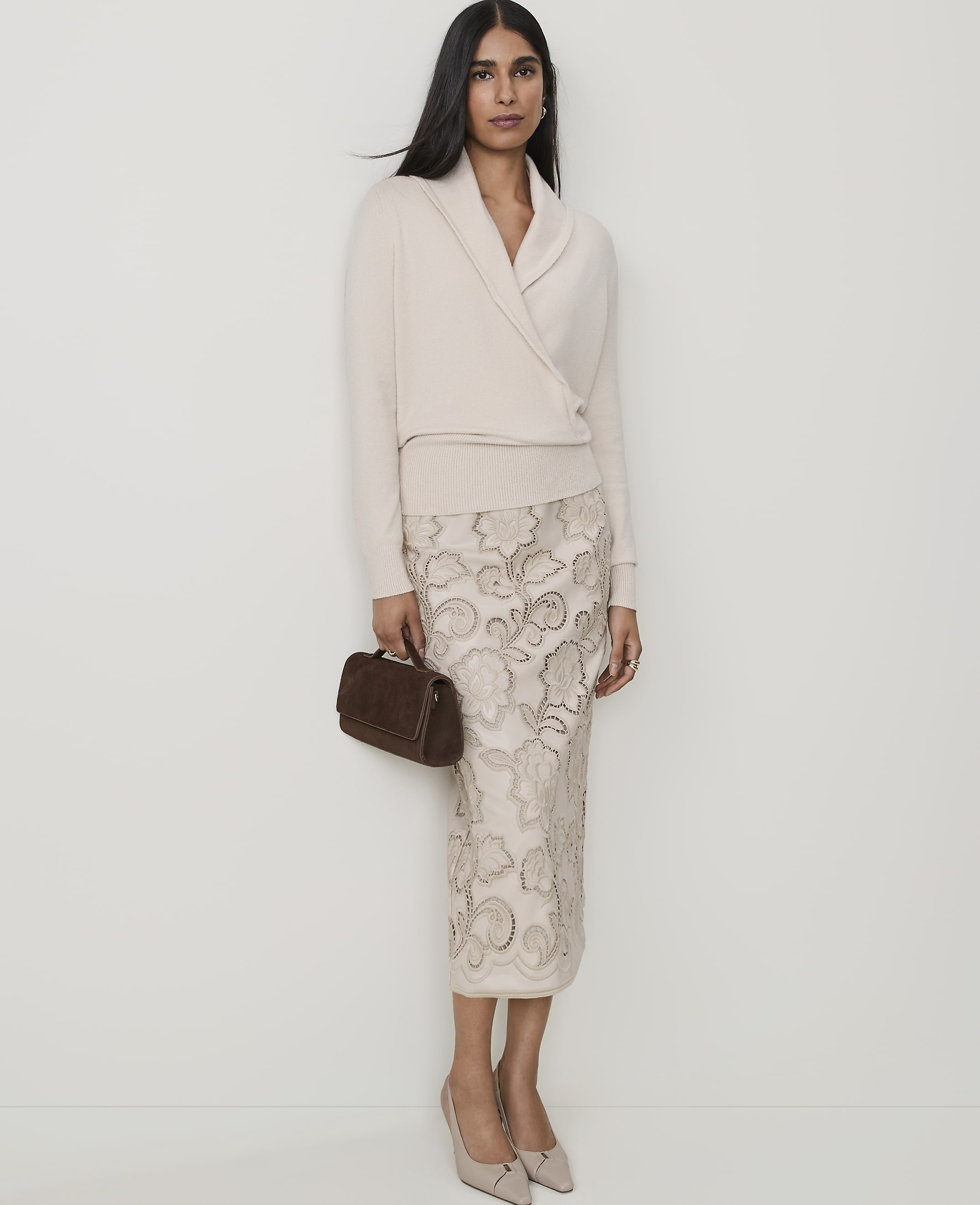 A woman in a cream wrap sweater and a patterned skirt stands against a plain white background, holding a small brown clutch.