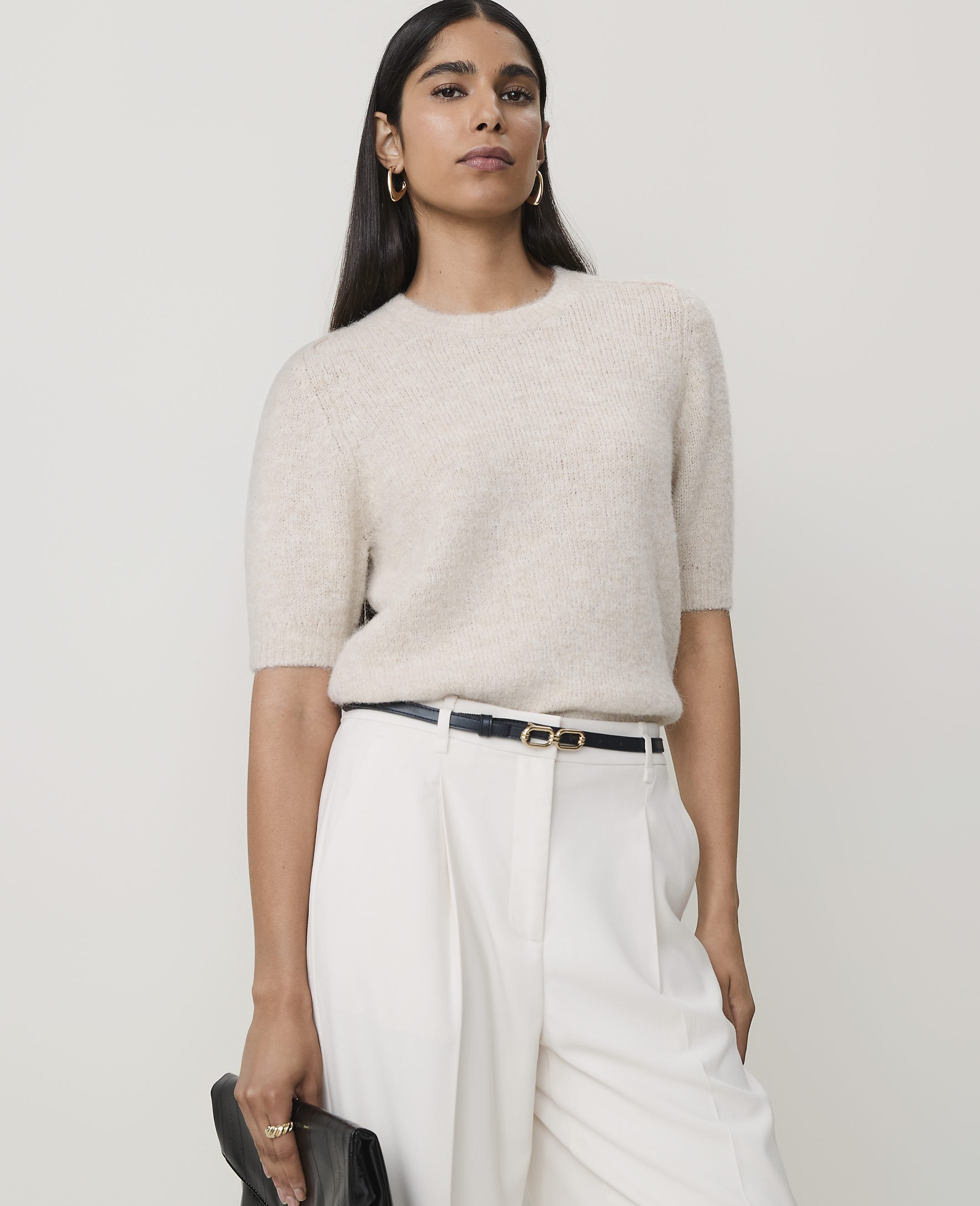 A woman in a cream sweater and white pants stands against a plain, light background, holding a black clutch.