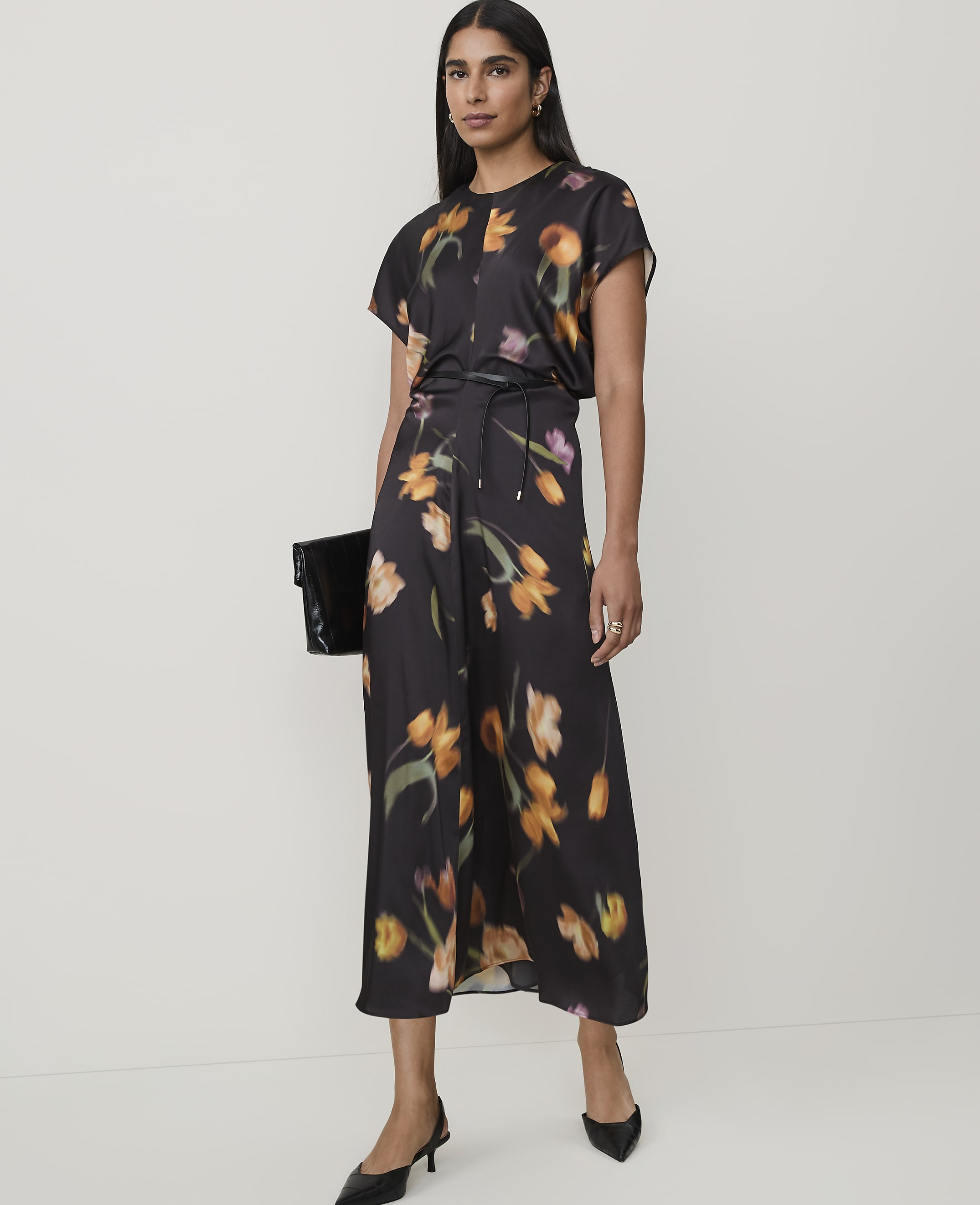 A woman in a floral dress and black heels stands against a plain background, holding a black clutch.