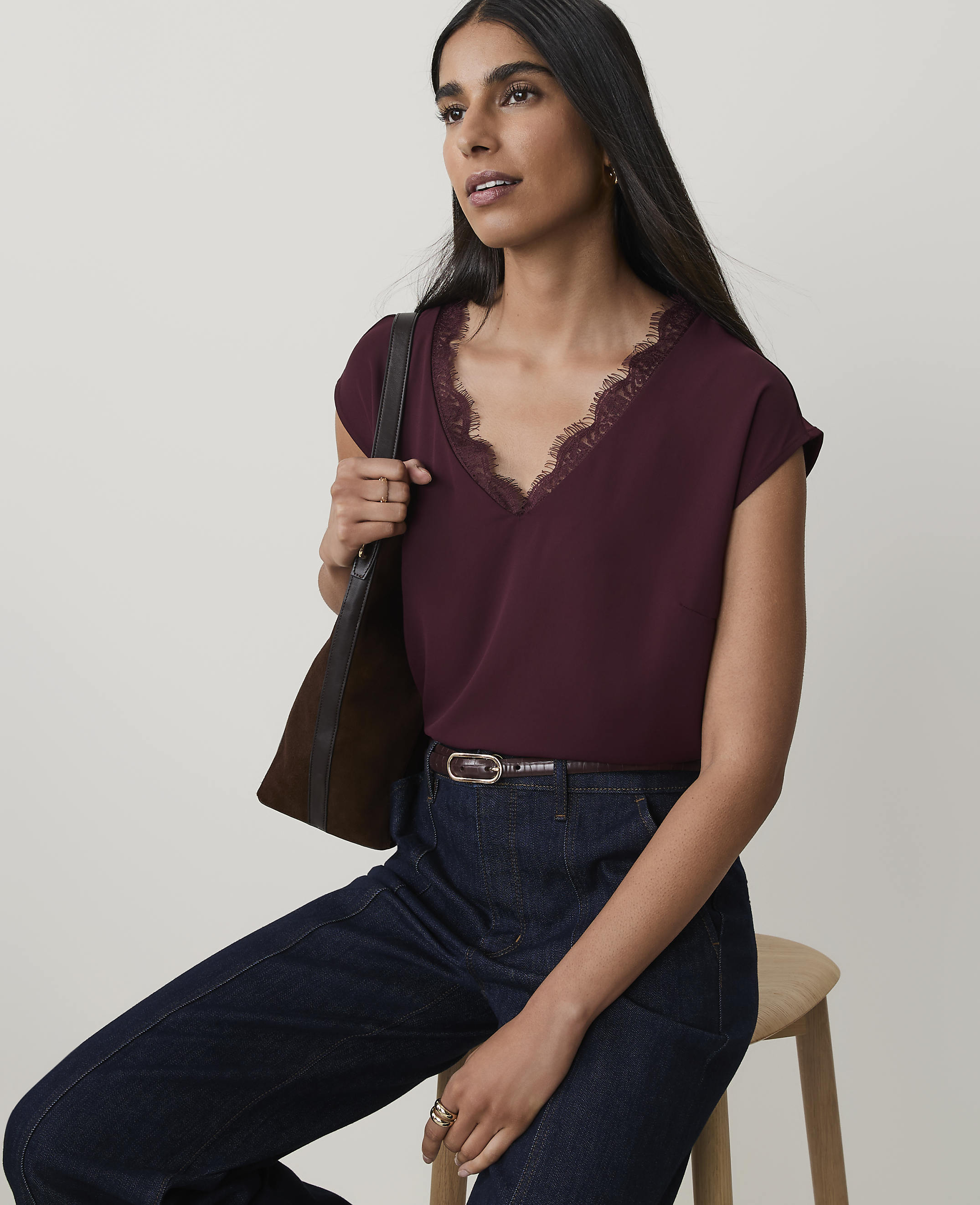 A woman in a burgundy lace-trimmed top and dark jeans sits against a plain, light background, carrying a brown bag over her shoulder.