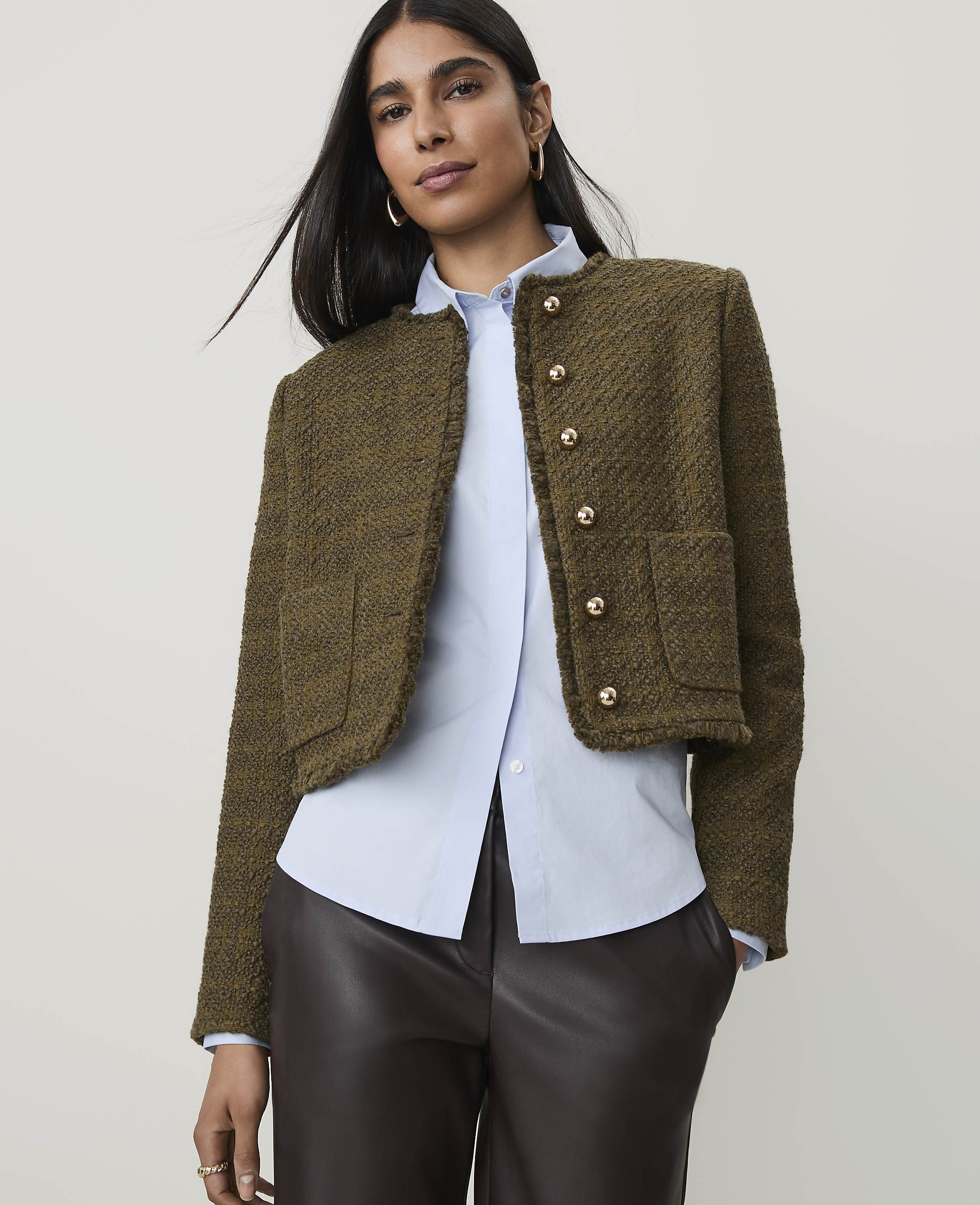 A woman in a textured olive jacket and light blue shirt stands against a plain background.