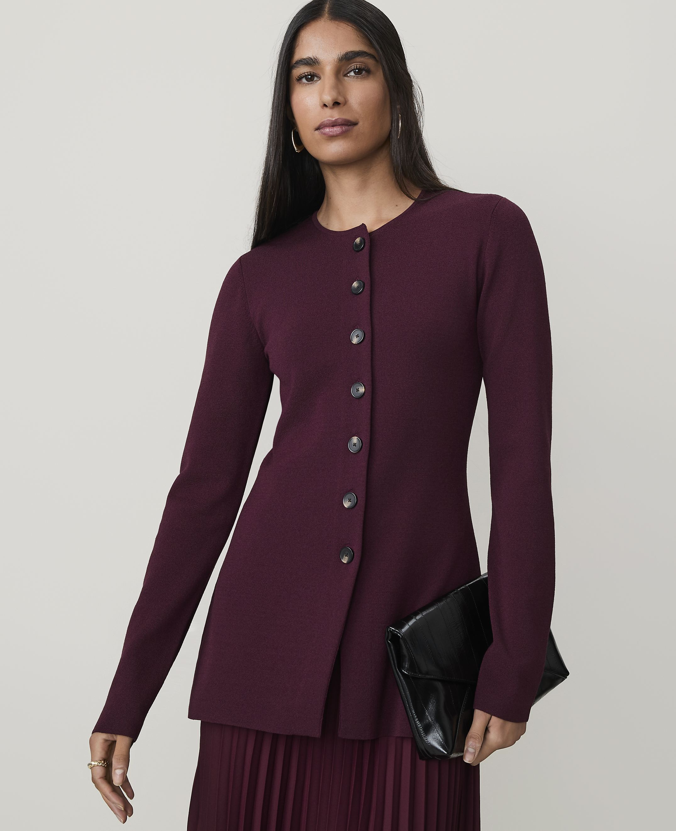 A woman in a burgundy cardigan and pleated skirt holds a black clutch against a plain background.