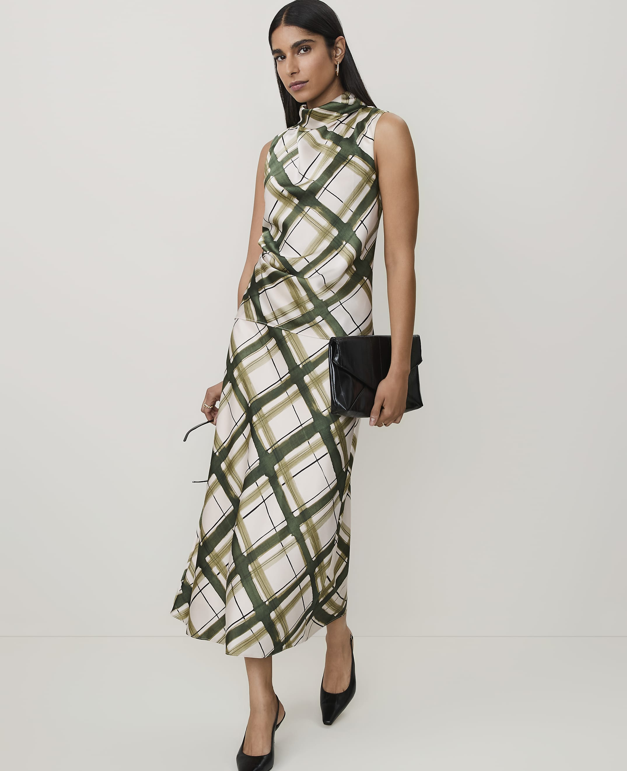 A woman in a plaid dress and black heels stands against a plain background, holding a black clutch and sunglasses.