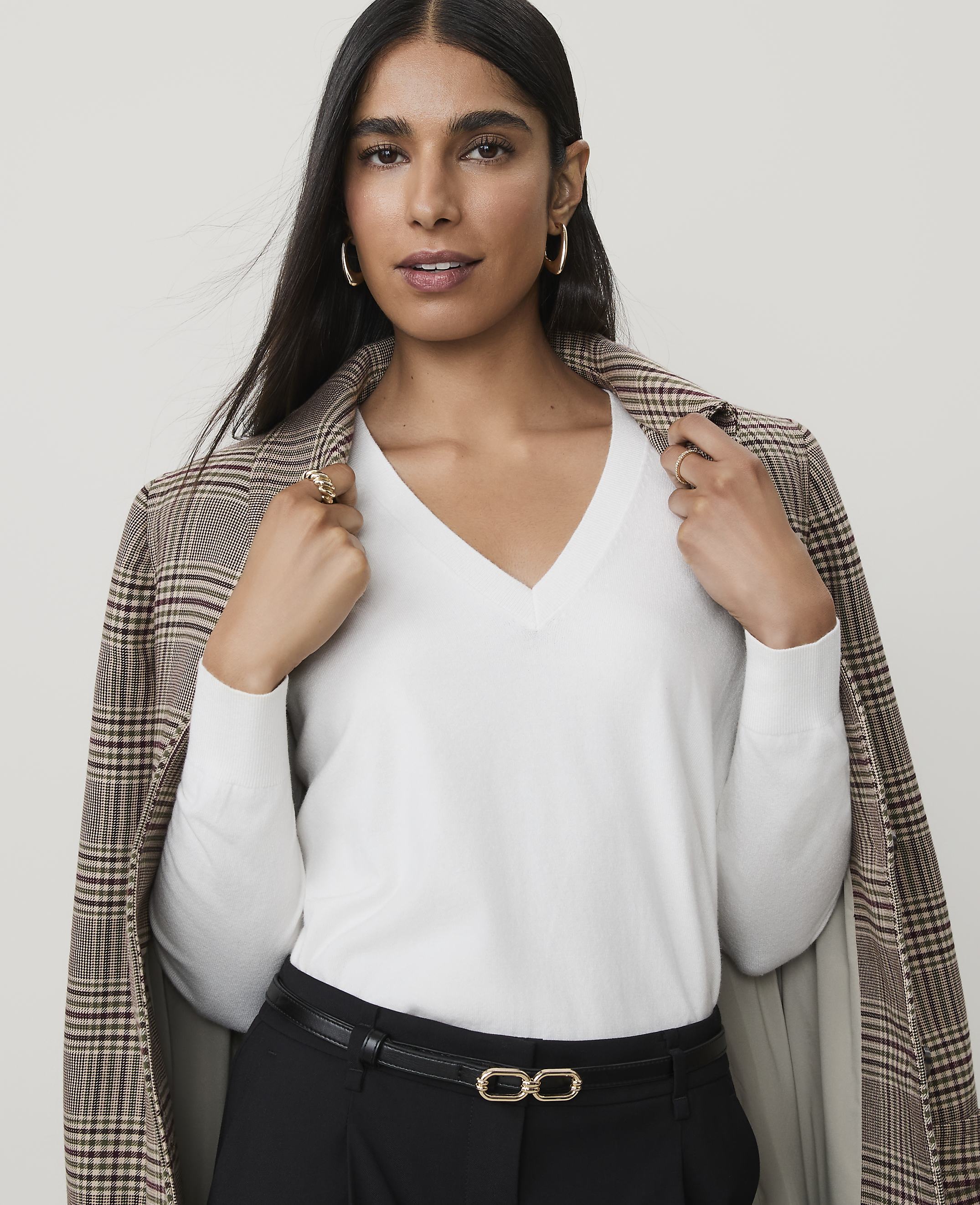 A woman in a white v-neck sweater and plaid blazer poses against a plain background.