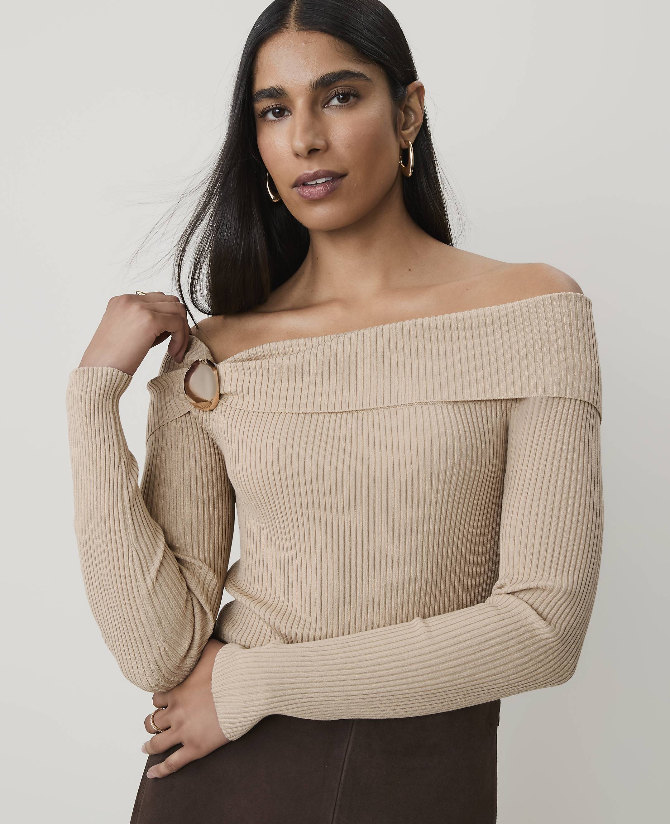 A woman in a beige off-the-shoulder sweater with a tortoiseshell ring detail poses against a plain, light gray background.