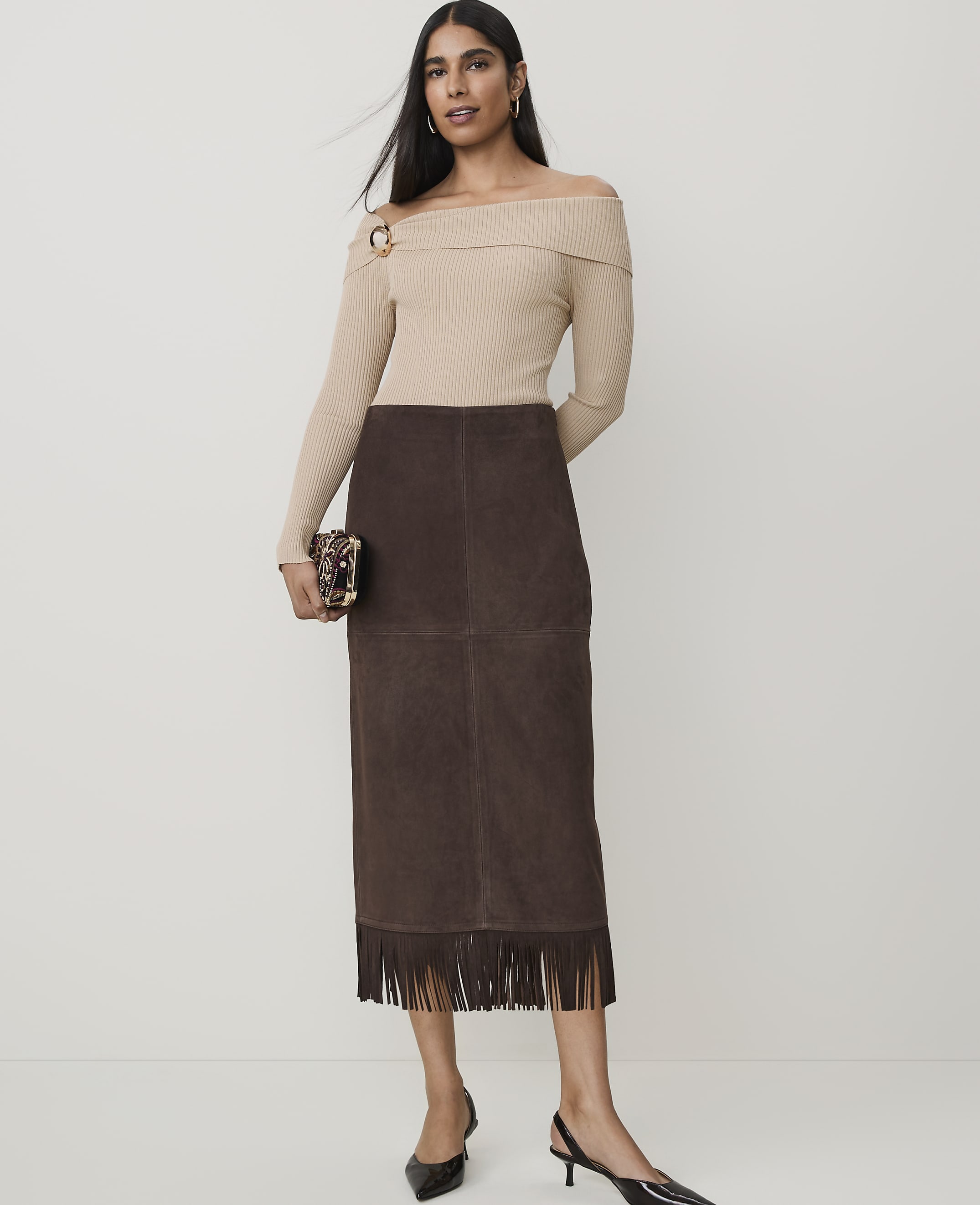 A woman in an off-the-shoulder sweater and suede fringe skirt stands against a plain white background, holding a patterned clutch.
