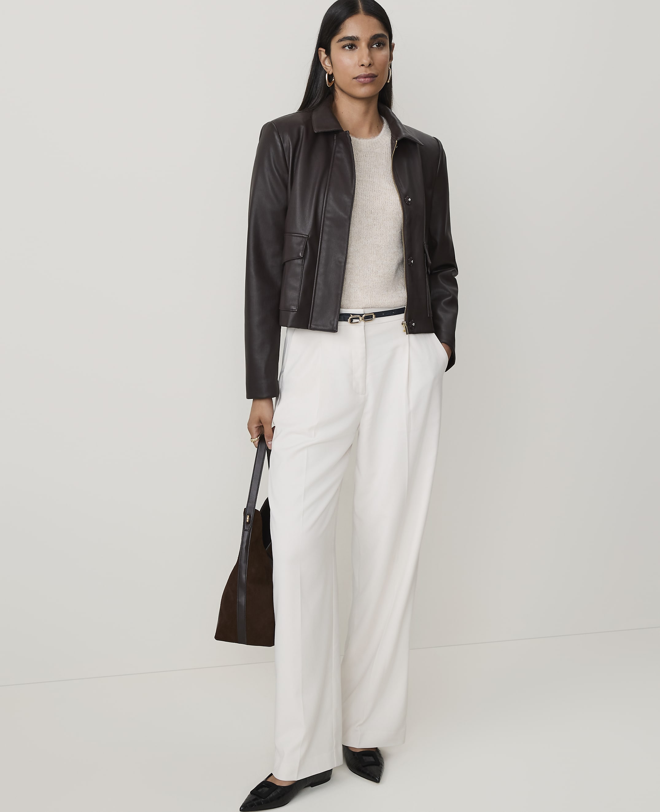 A woman in a cream sweater, dark brown leather jacket, and white wide-leg pants stands against a plain white background, holding a dark brown tote bag.