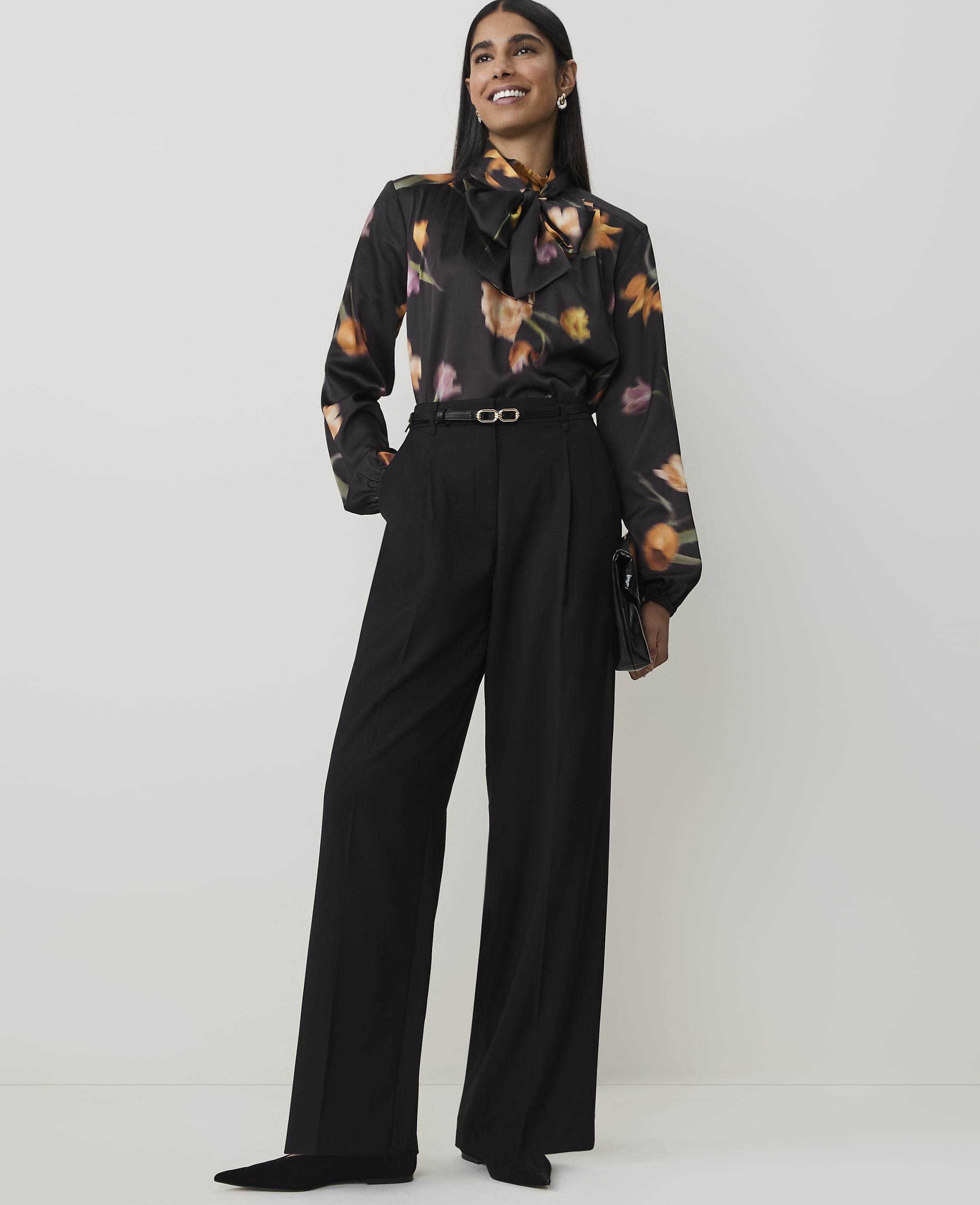 A woman in a floral blouse and black wide-leg pants stands against a plain, light background.
