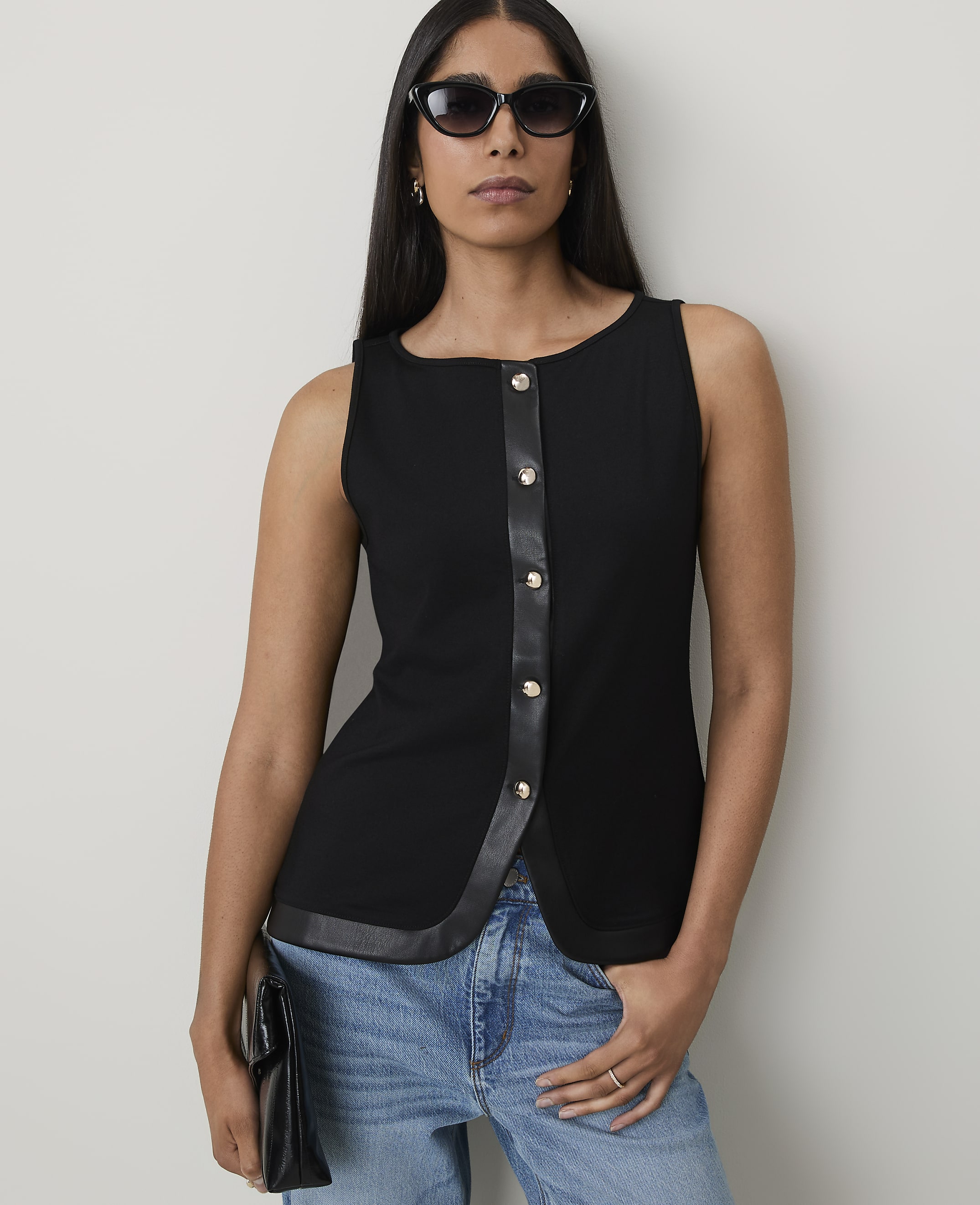 A woman in a black sleeveless top and jeans stands against a plain background, holding a black clutch.