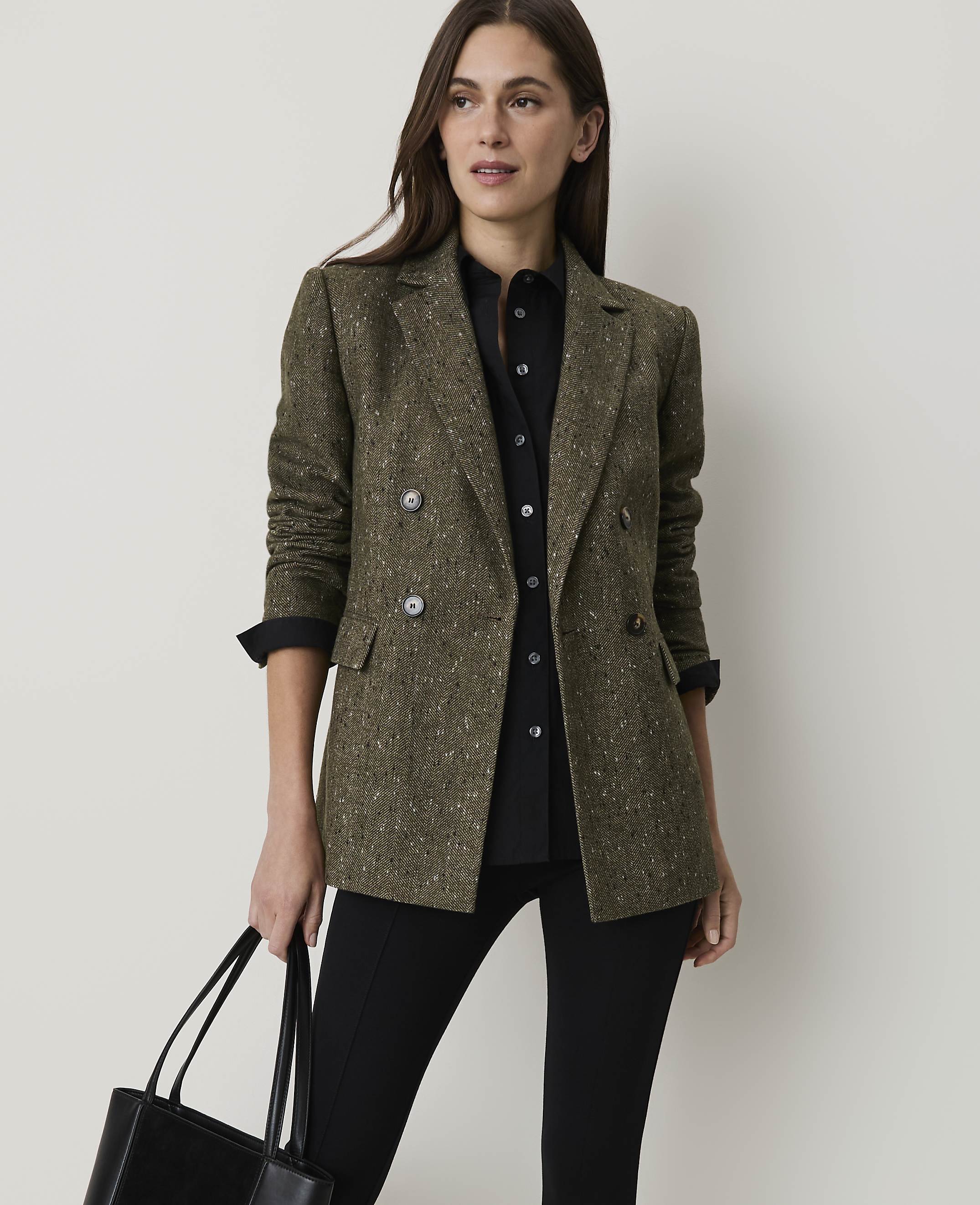A woman in a tweed blazer and black shirt holds a tote bag in front of a plain background.
