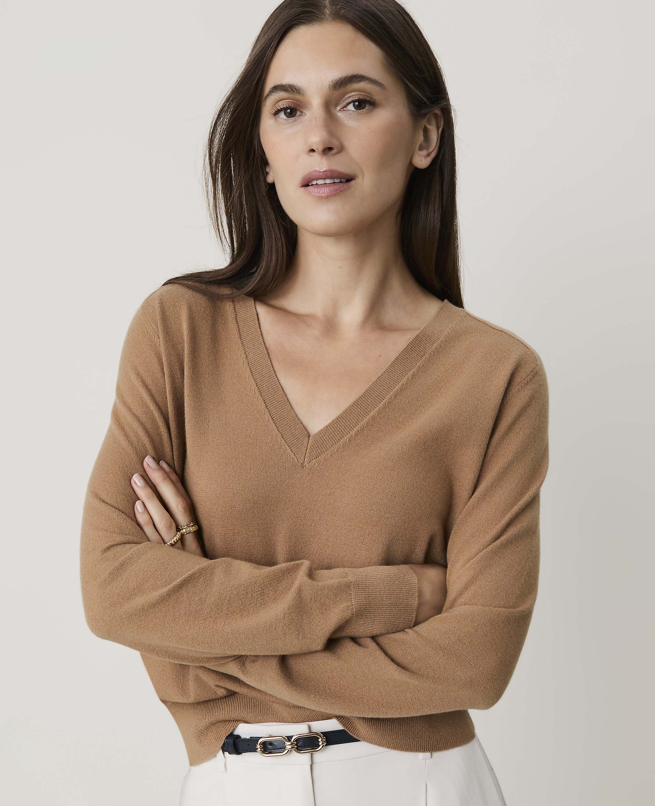 A woman in a tan v-neck sweater stands with her arms crossed against a plain white background.