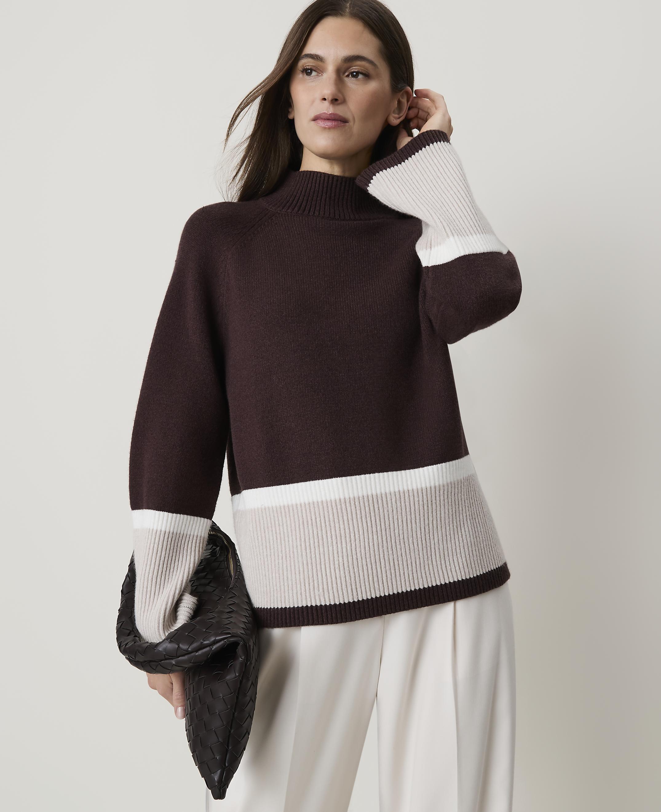 A woman in a burgundy sweater with cream and beige stripes holds a dark clutch in front of a plain background.