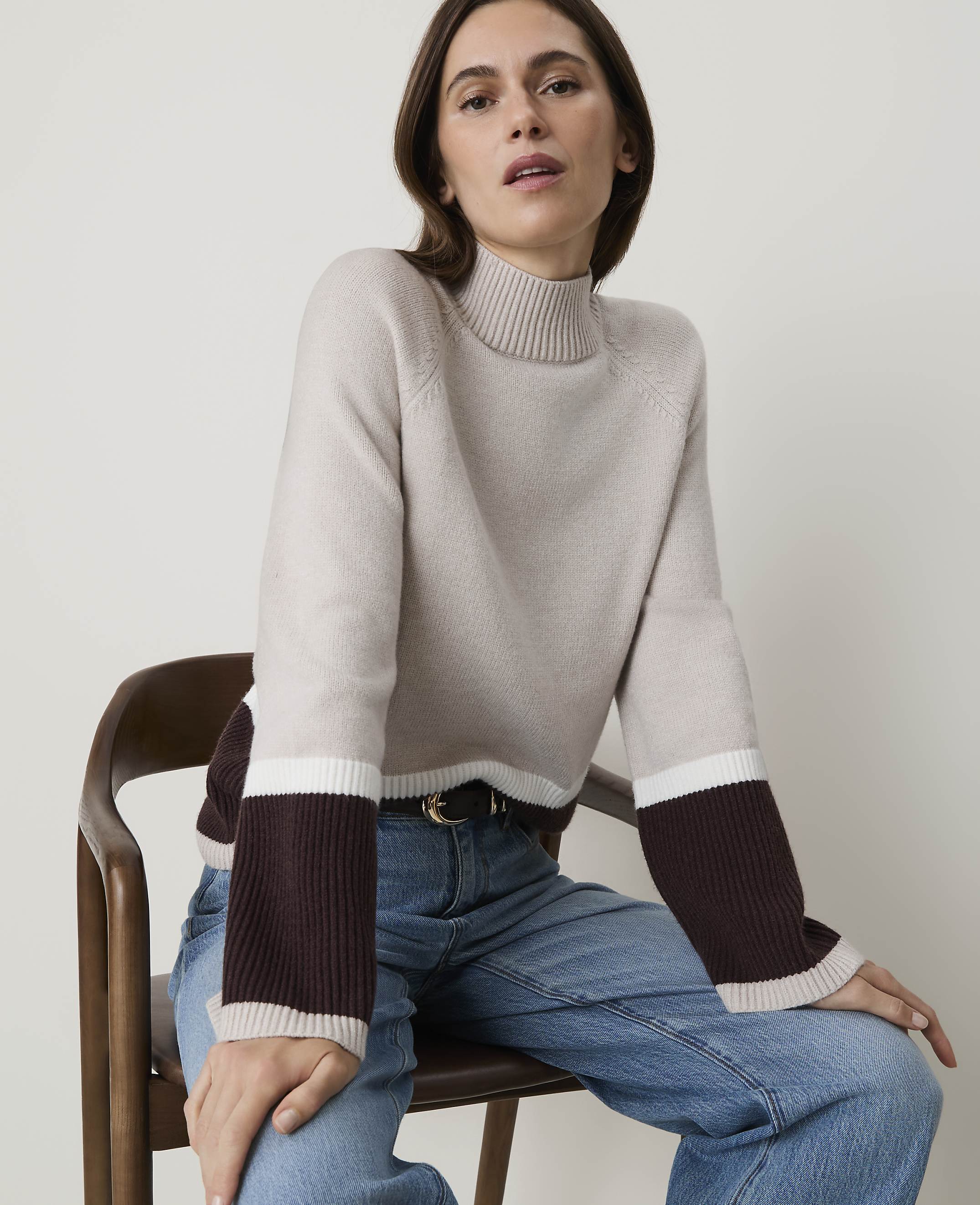 A woman in a color-blocked sweater and jeans sits on a wooden chair against a plain background.