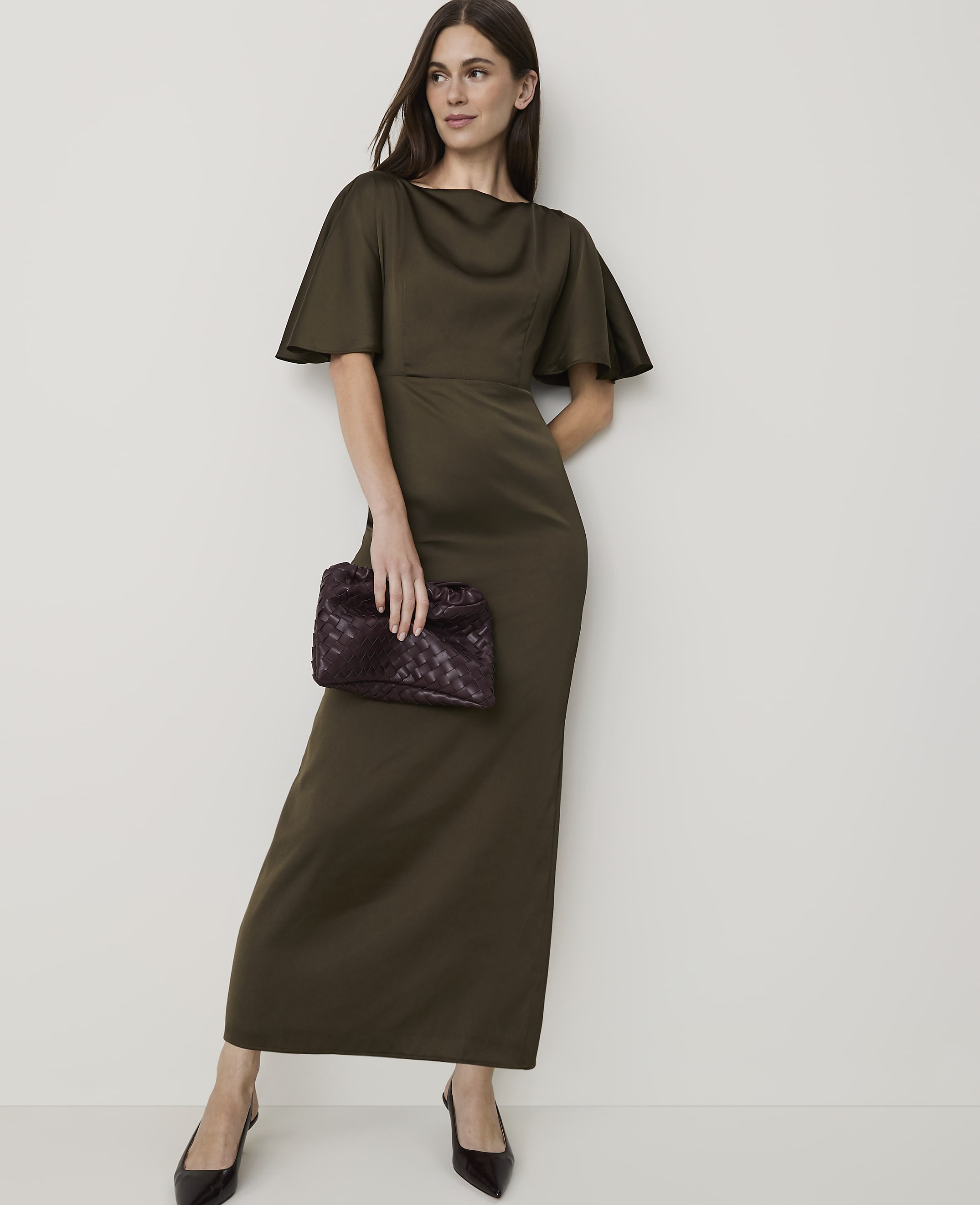 A woman in a long, olive-green dress and dark heels holds a patterned clutch in front of a plain, off-white background.