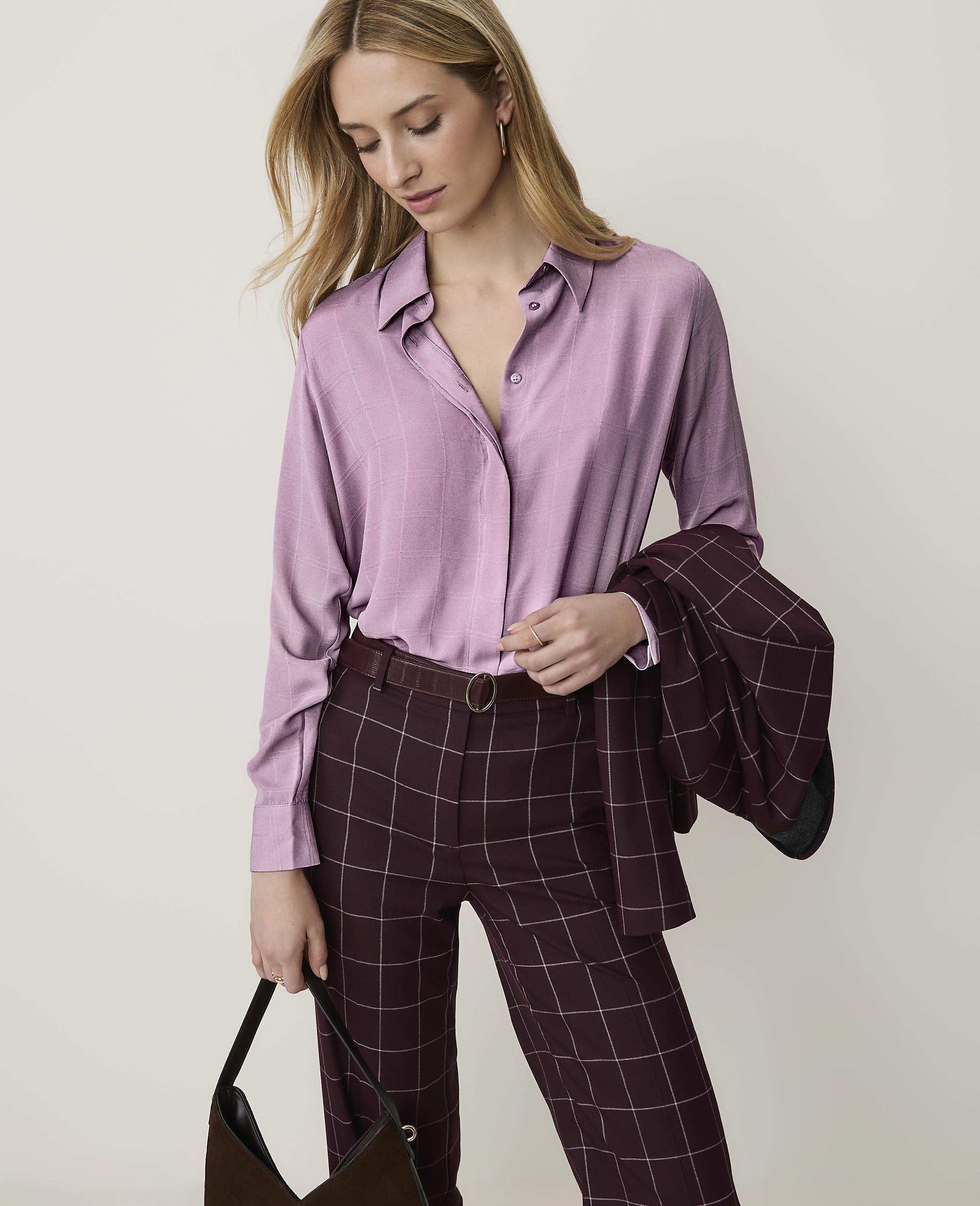 A woman in a lilac blouse and plaid pants stands against a plain white background, holding a dark handbag.