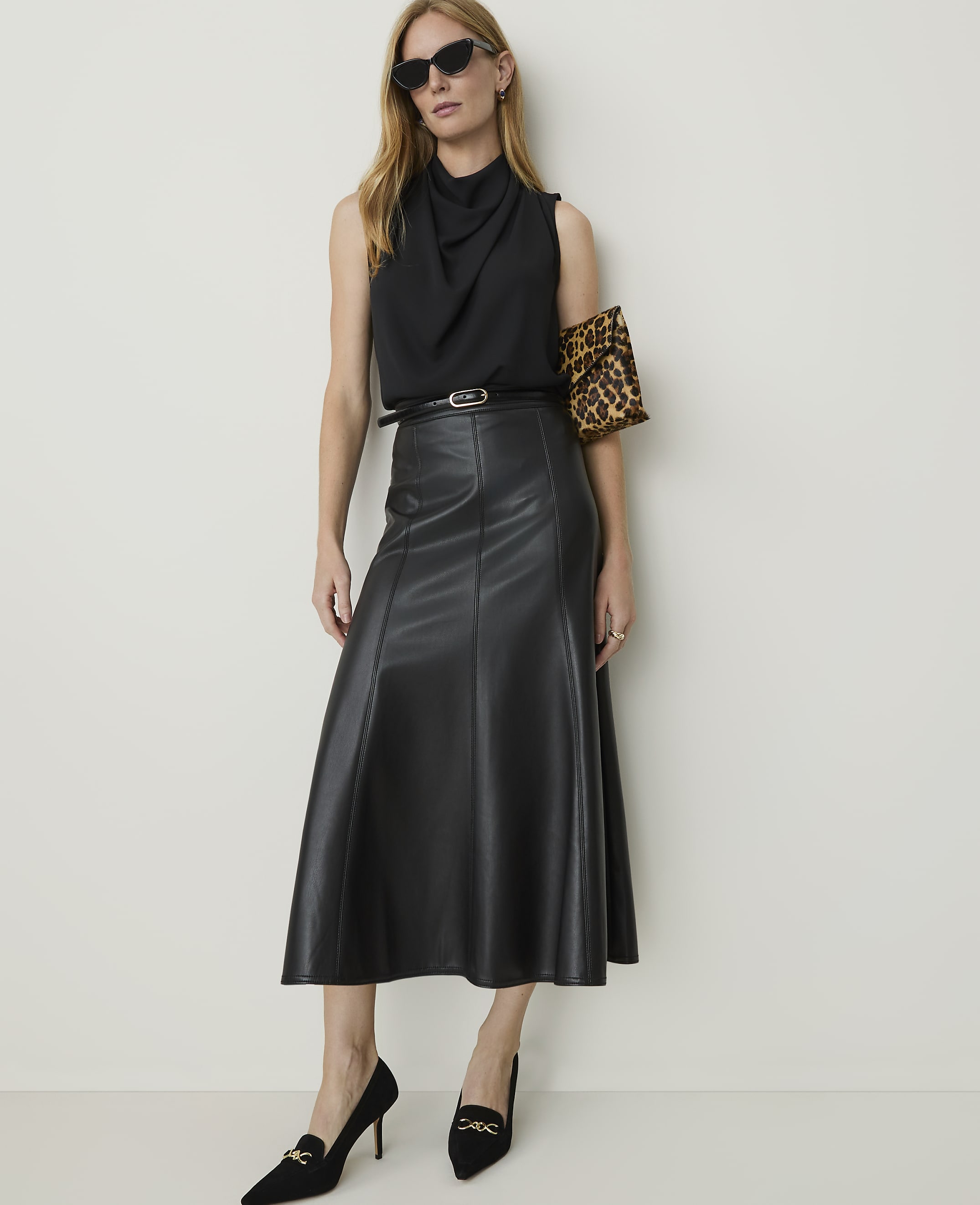 A woman in a black cowl-neck top and a black leather midi skirt poses against a plain white background, accessorized with sunglasses, a leopard-print clutch, and black heels.