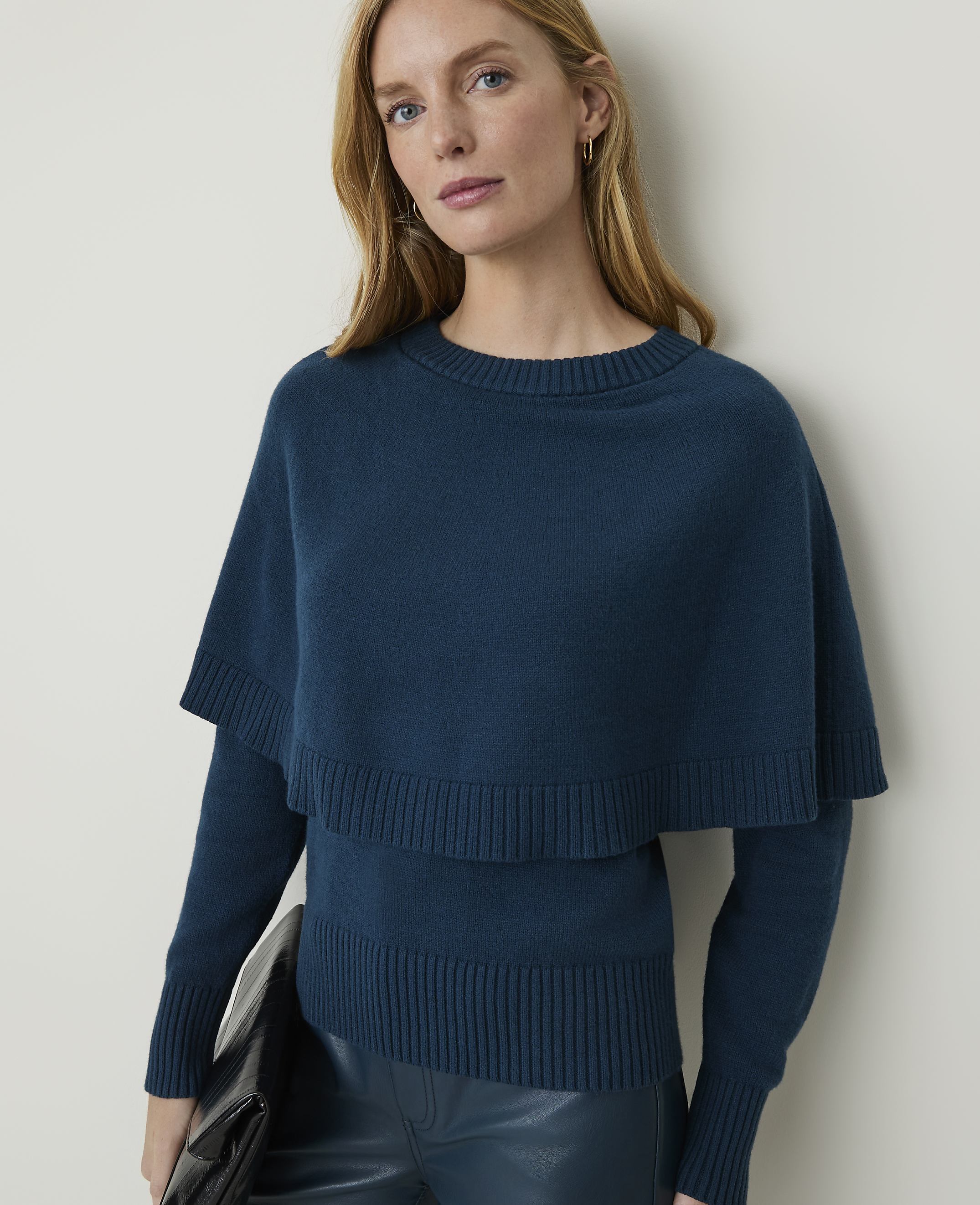 A woman in a teal sweater with a capelet and dark pants holds a black clutch against a plain background.