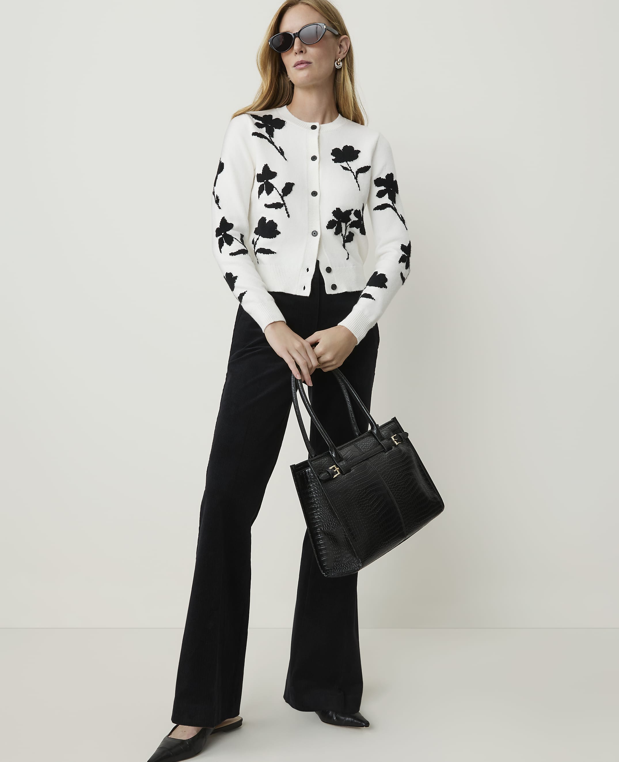 A woman in a floral cardigan and black flared pants poses with a black handbag against a plain white background.