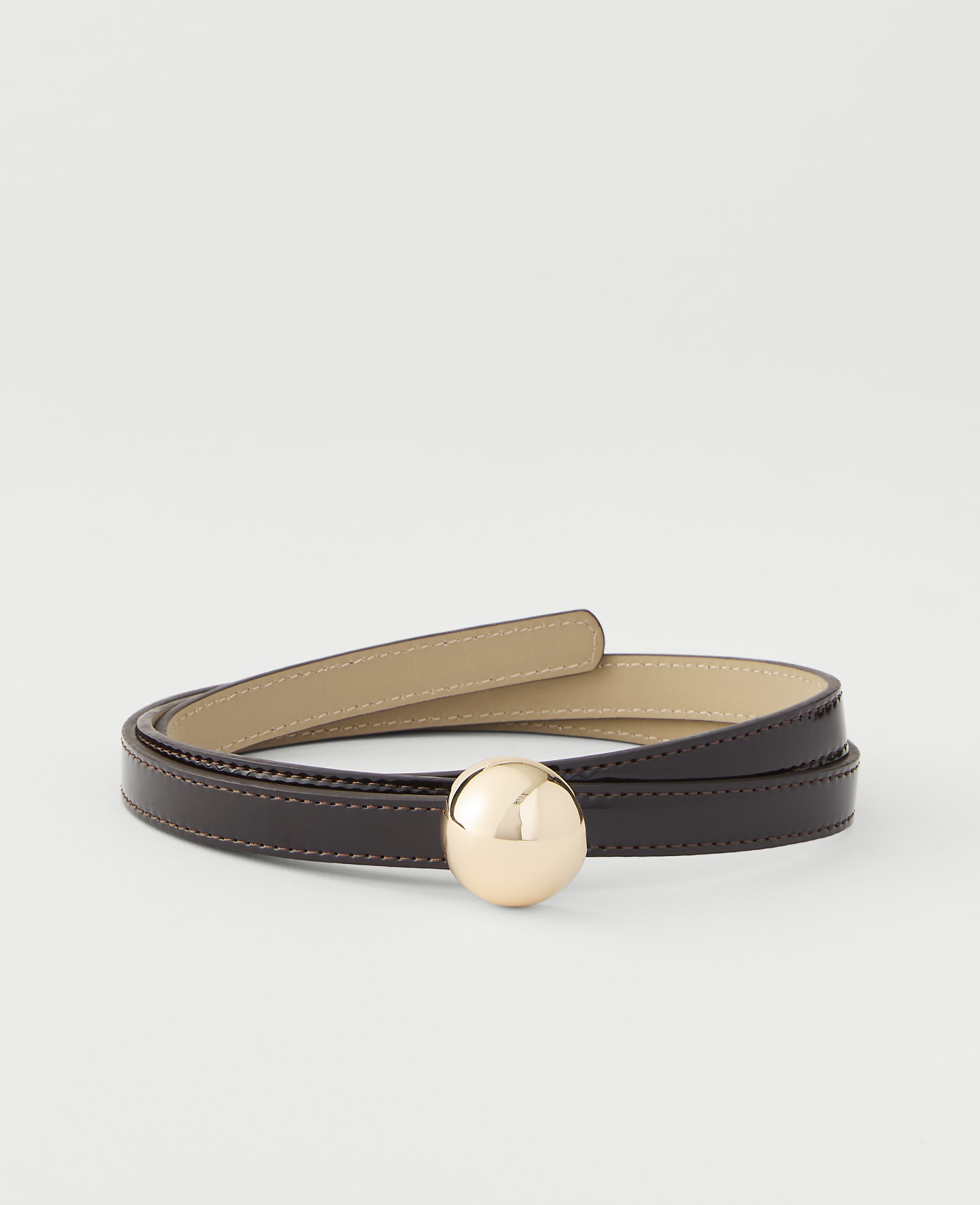 A thin, dark brown leather belt with a gold sphere buckle is coiled on a plain white background.