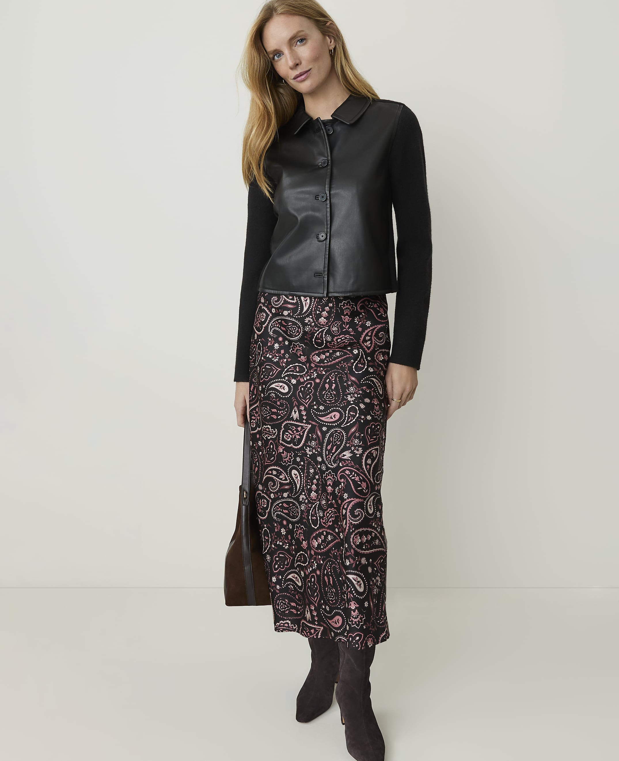 A woman in a black leather jacket and a patterned skirt stands against a plain white background, holding a brown handbag.