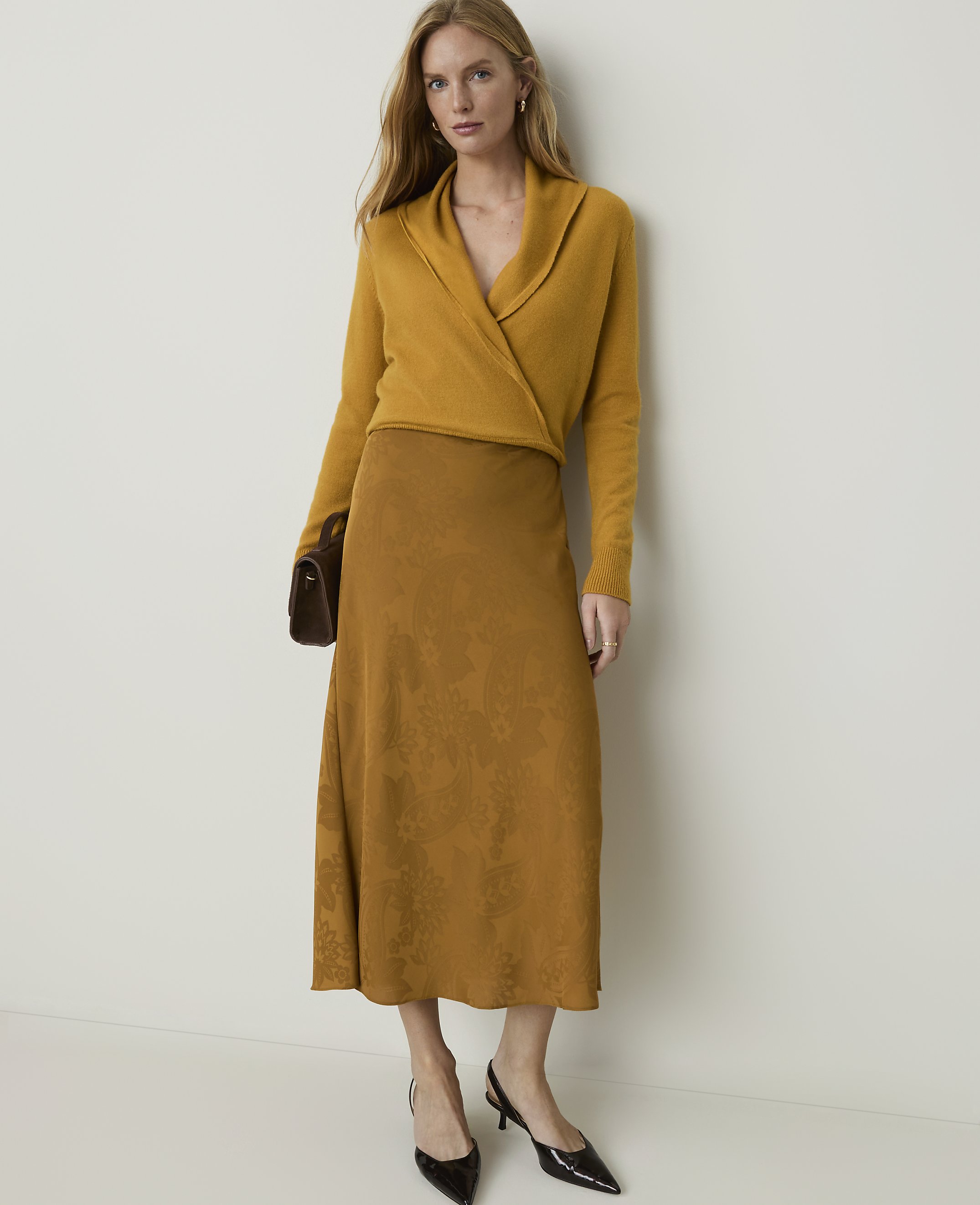 A woman in a mustard yellow wrap sweater and a patterned brown skirt stands against a plain cream background, holding a dark brown clutch.