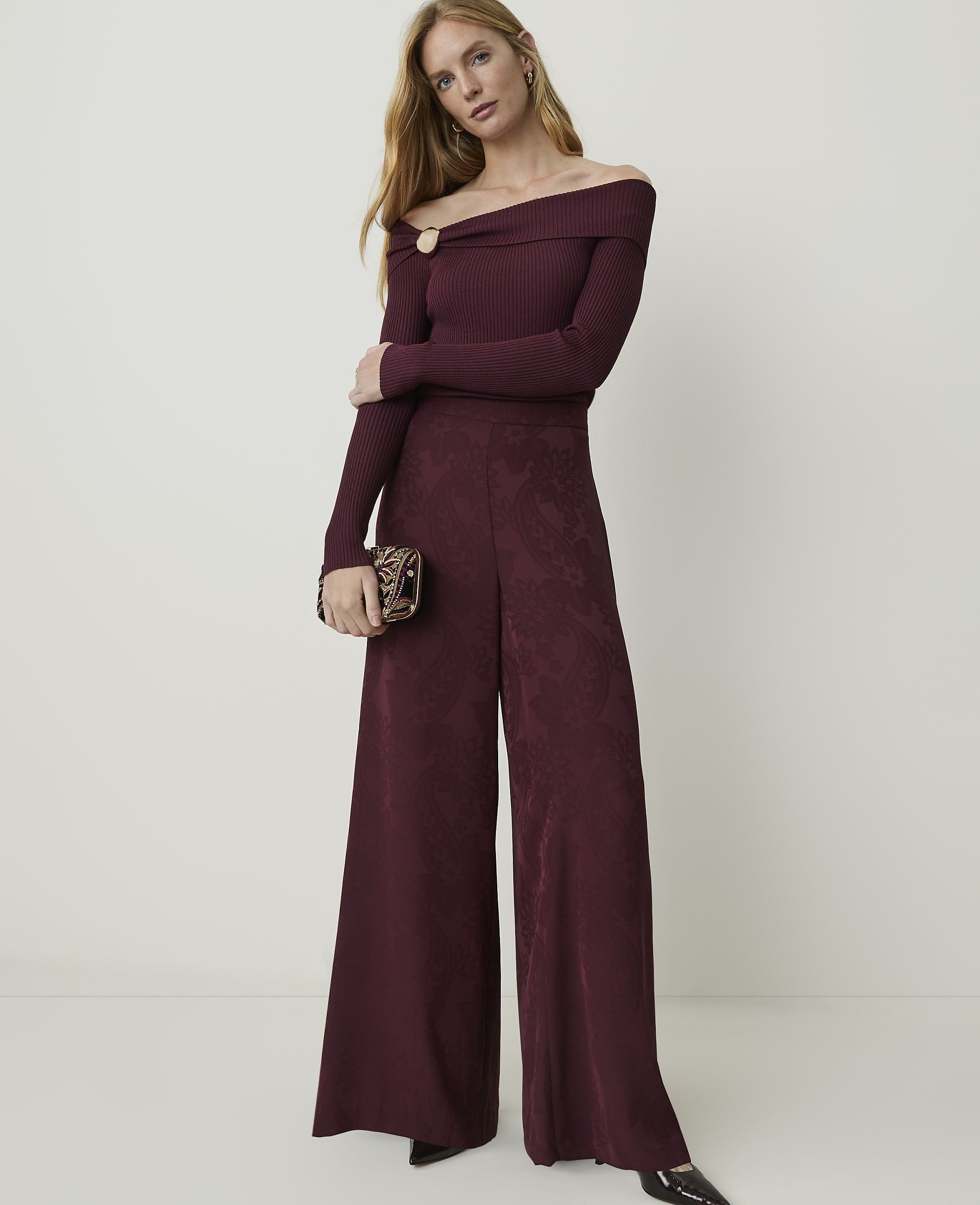 A woman in a burgundy off-the-shoulder jumpsuit and patterned clutch stands against a plain, light-colored background.