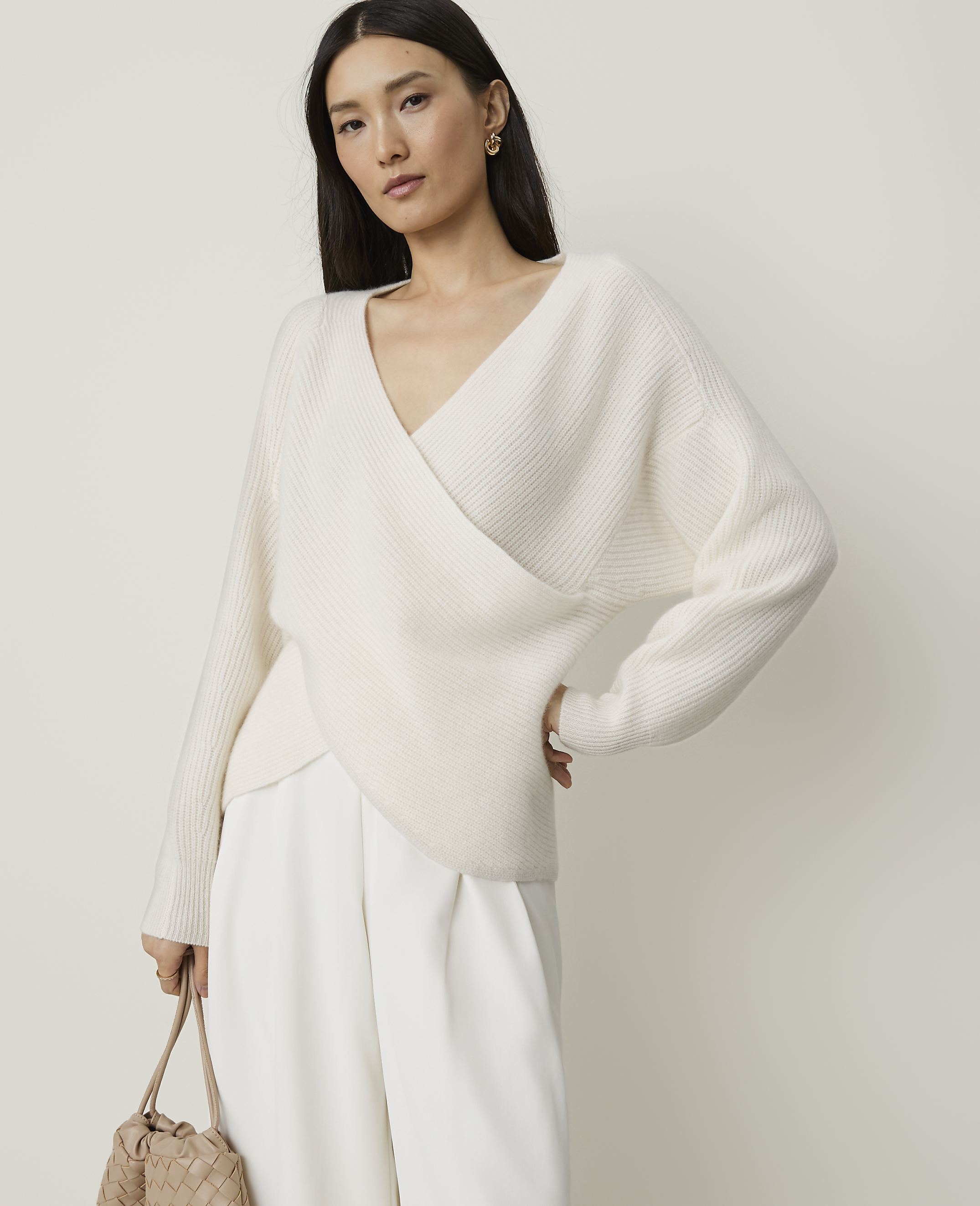 A woman in a cream wrap sweater and white pants stands against a plain, light background, holding a woven bag.