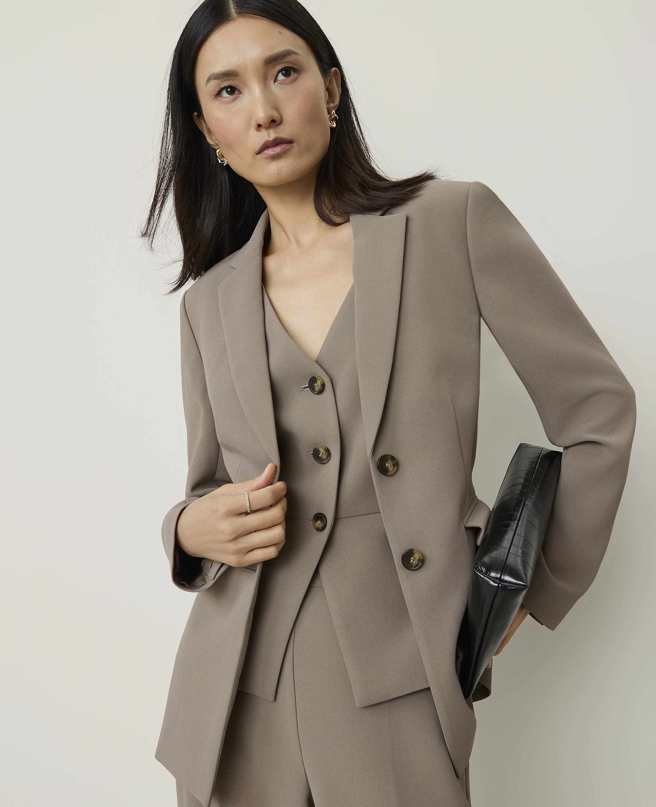 A woman in a taupe suit and holding a black clutch stands against a plain, light background.