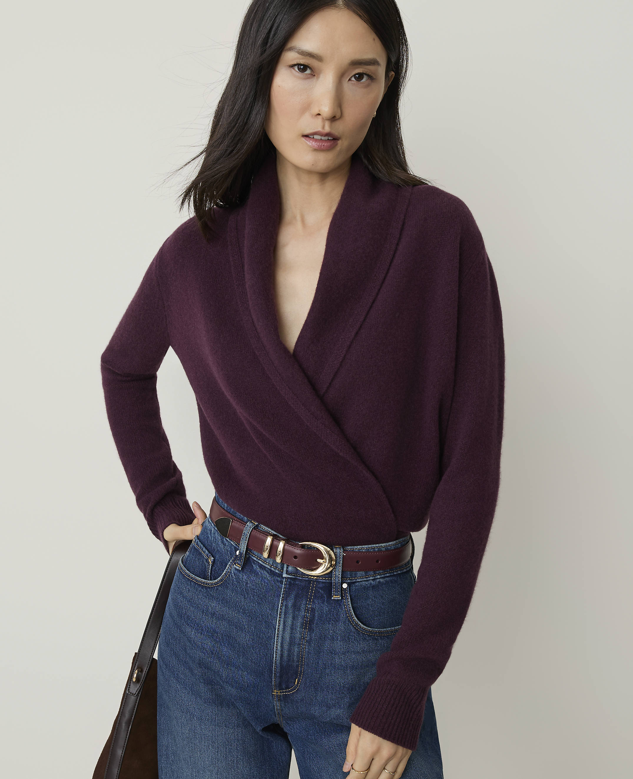 A woman in a plum-colored wrap sweater and blue jeans stands against a plain, light gray background.